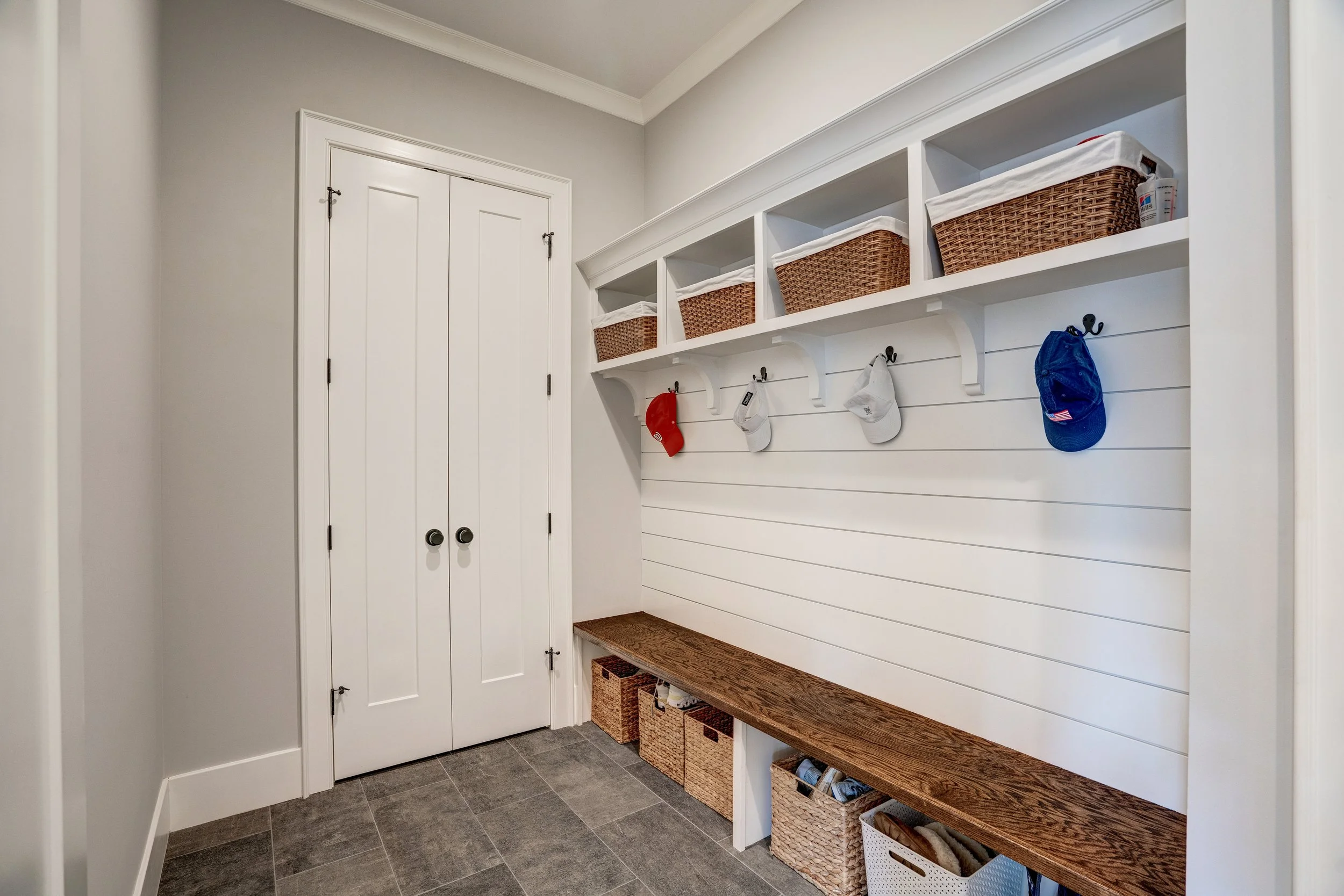A mudroom with white walls, dark tile floor, double white doors, and built-in shelves with baskets and hooks holding hats.