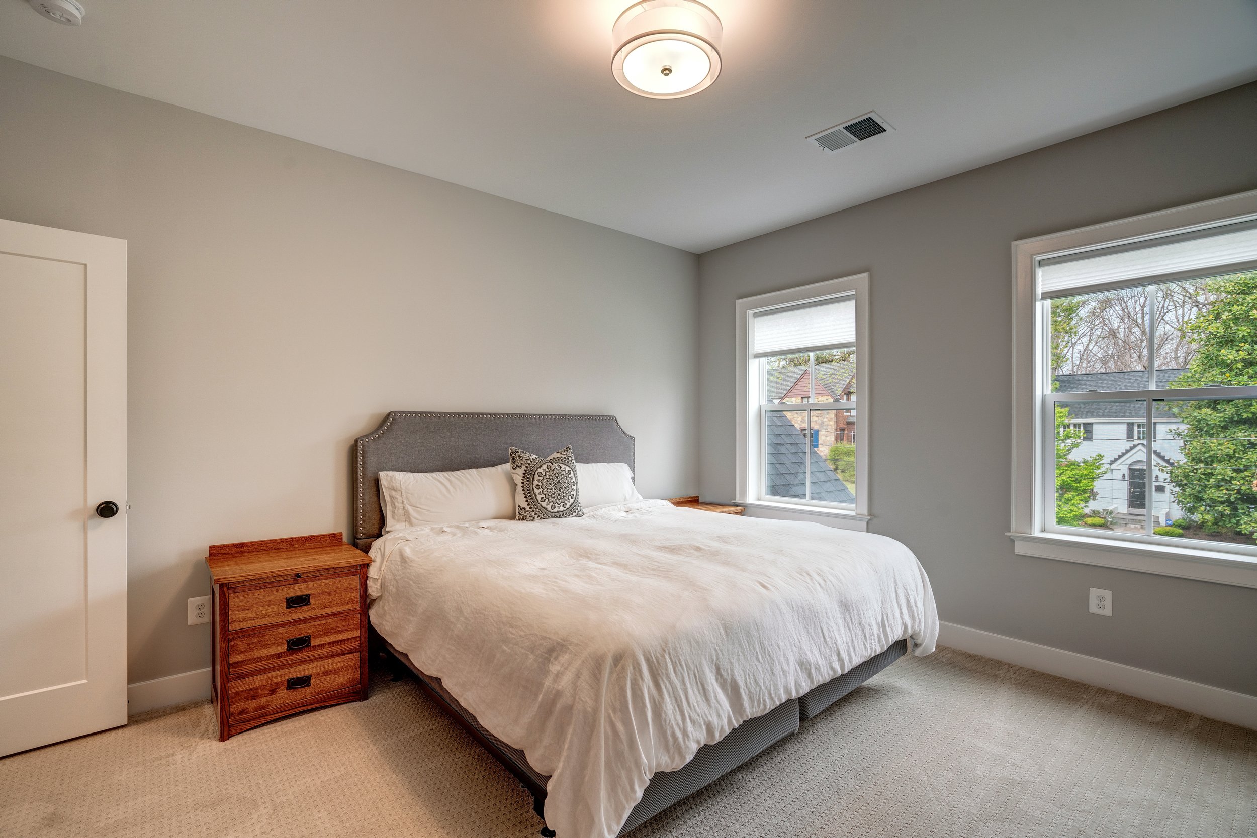 A bedroom with a bed, two windows, a wooden nightstand, and a ceiling light.