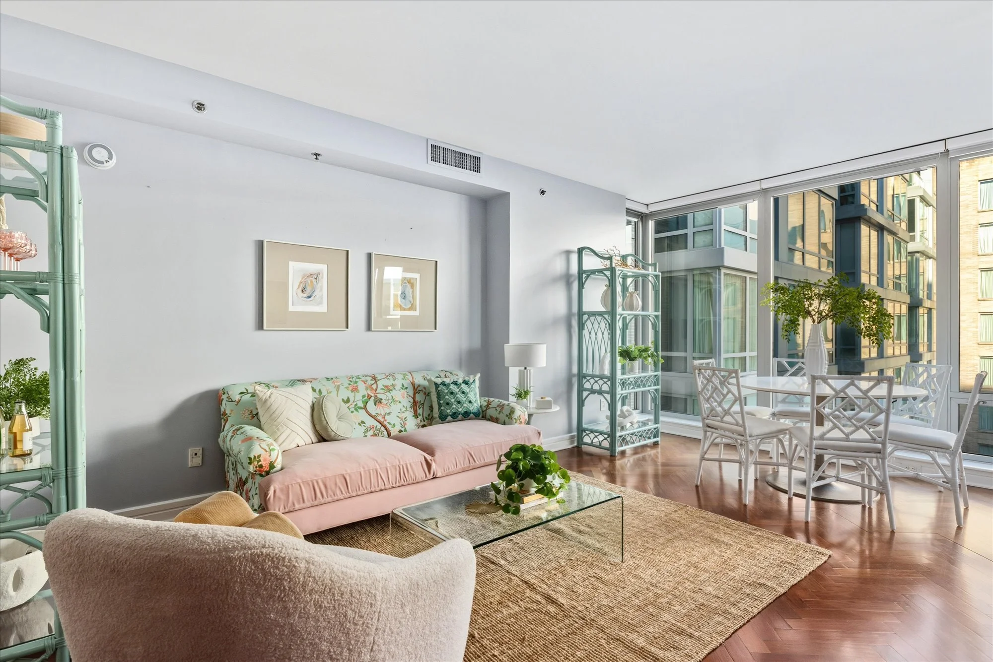 Living room with pink floral sofa, white chairs around a table, green plants, artwork on a light wall, and large windows with city views.