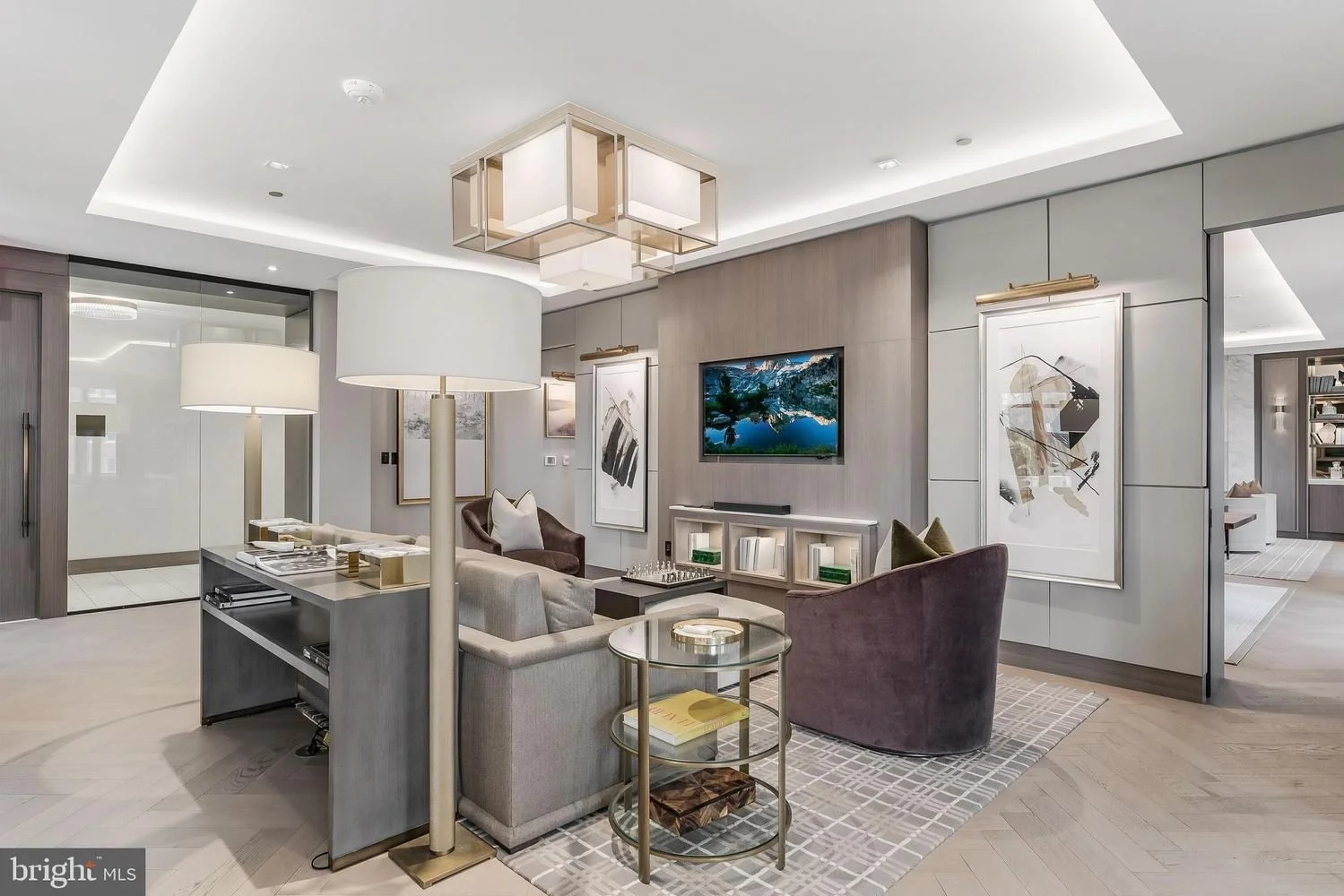 Modern living room with beige and gray furniture, wall-mounted TV, abstract artwork, glass side table, and contemporary lighting fixtures.