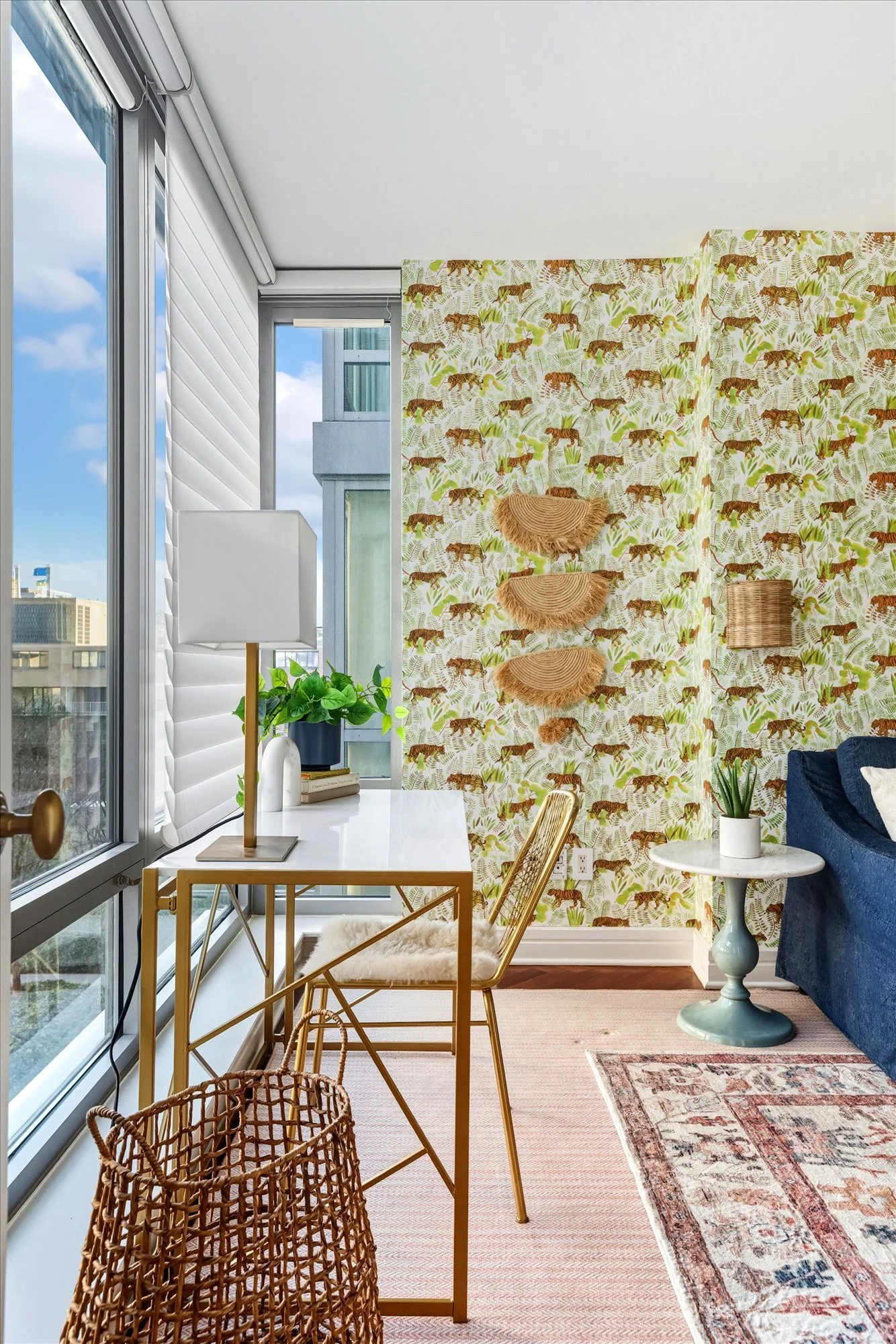 Sunlit indoor space with large windows, a white table with gold frame, wicker basket, amber chairs, printed wallpaper with tigers and tropical leaves, potted plants, a blue sofa, and a patterned rug.
