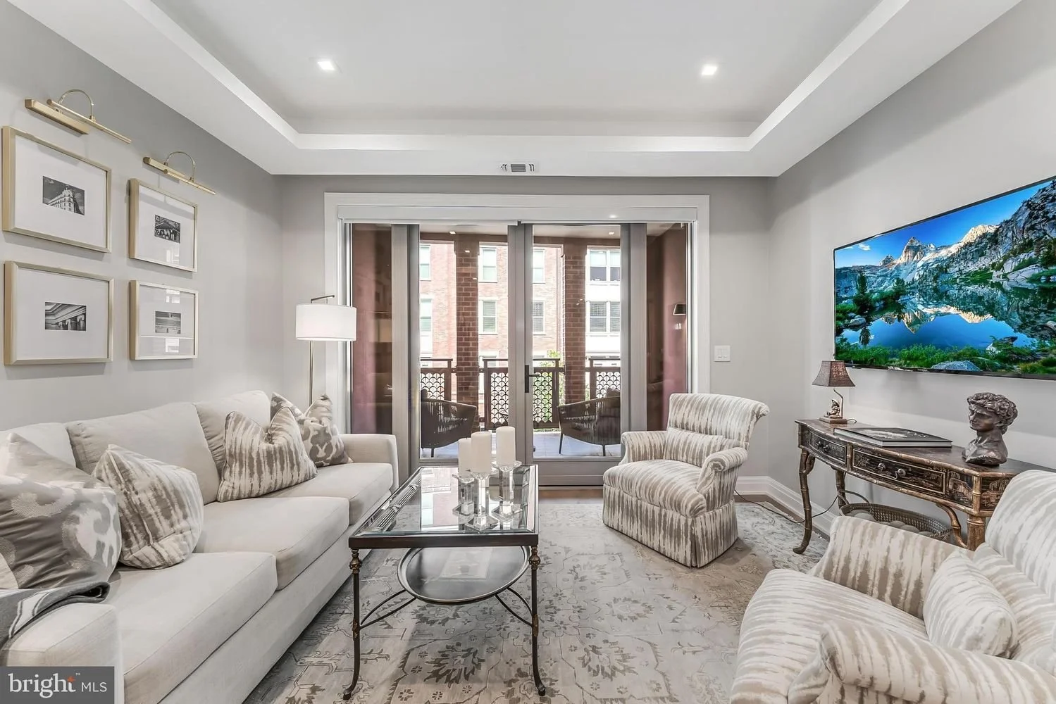 A modern living room with light gray walls, white ceiling, and a sliding glass door leading to a balcony. The room has a cream-colored sofa with multiple patterned pillows, a glass-top coffee table, a patterned armchair, and a vintage-style side tabl