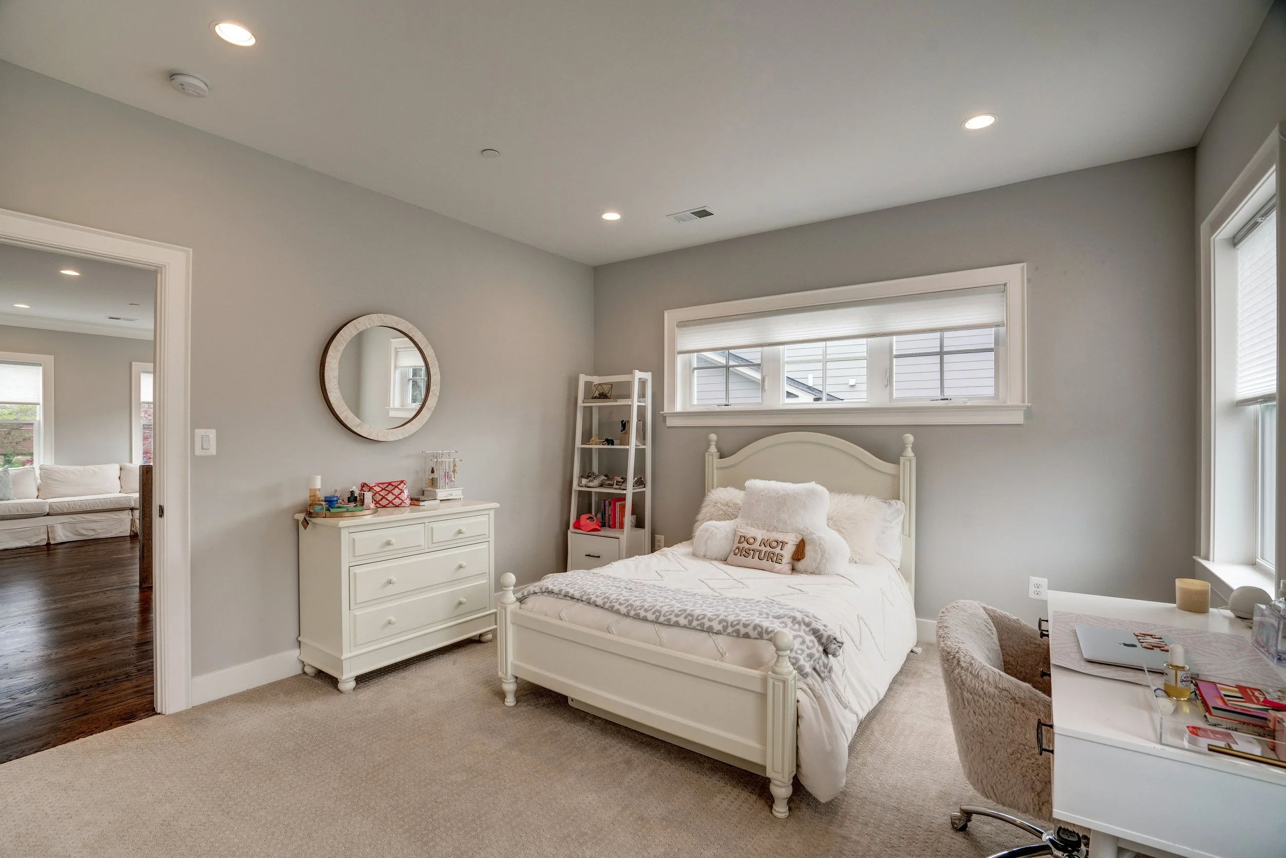 A cozy bedroom with a white bed, plush pillows including one with the words 'Do Not Disturb,' a white dresser, a round mirror on the wall, a window with blinds, a ladder shelf with decorative items, and a carpeted floor.