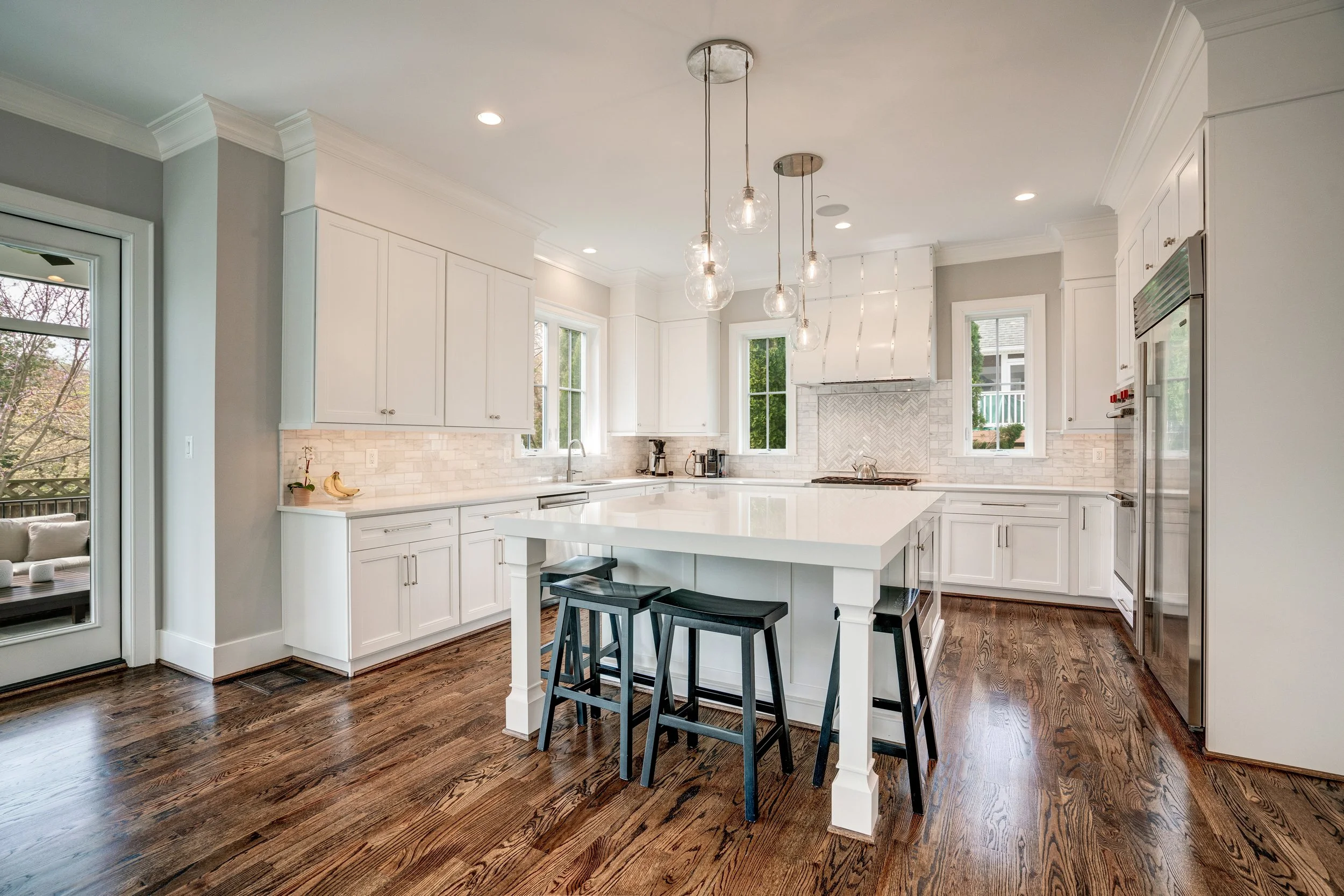 Modern white kitchen with island, pendant lights, dark wood floors, and bright windows.