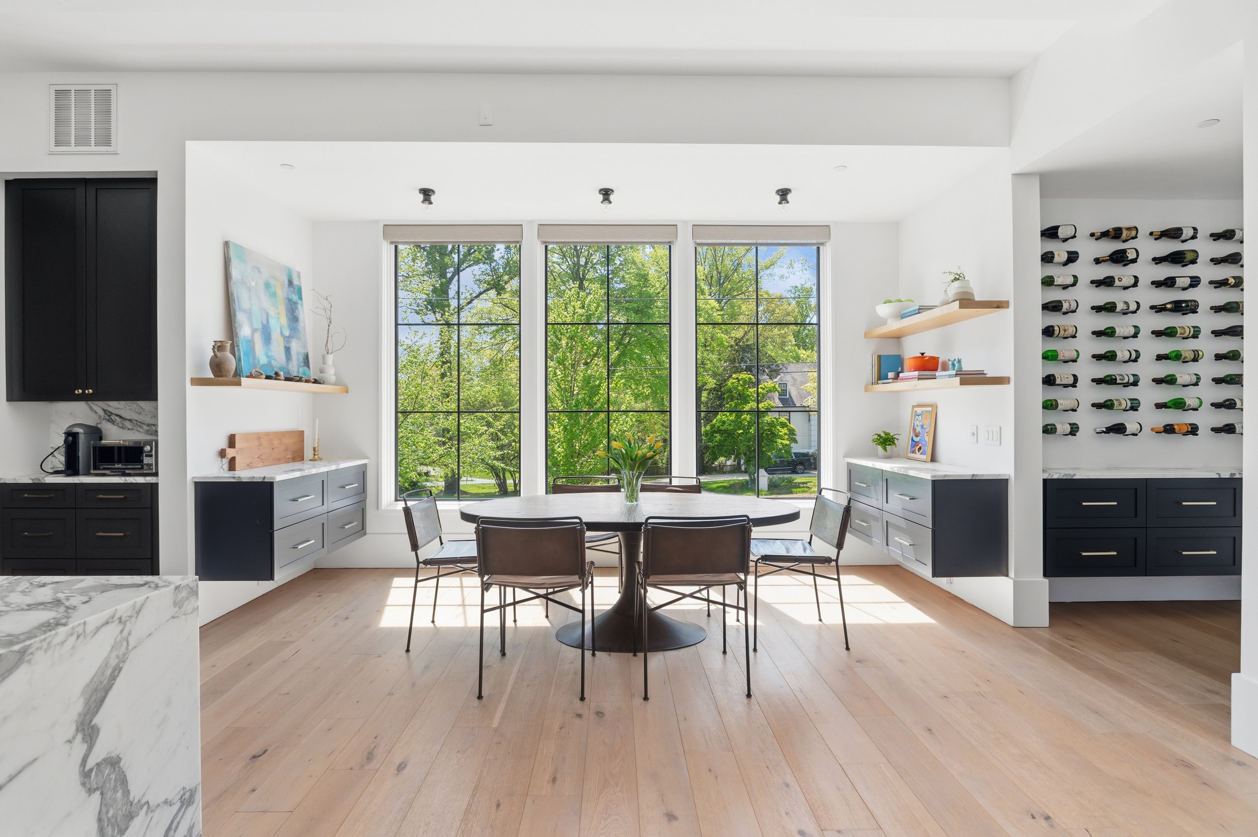 Bright dining area with large window showing green trees, a round table, and four chairs. Shelves and a wine rack are on the white walls, with decorative items and books. Sunlight streams through the window, illuminating the wooden floor.