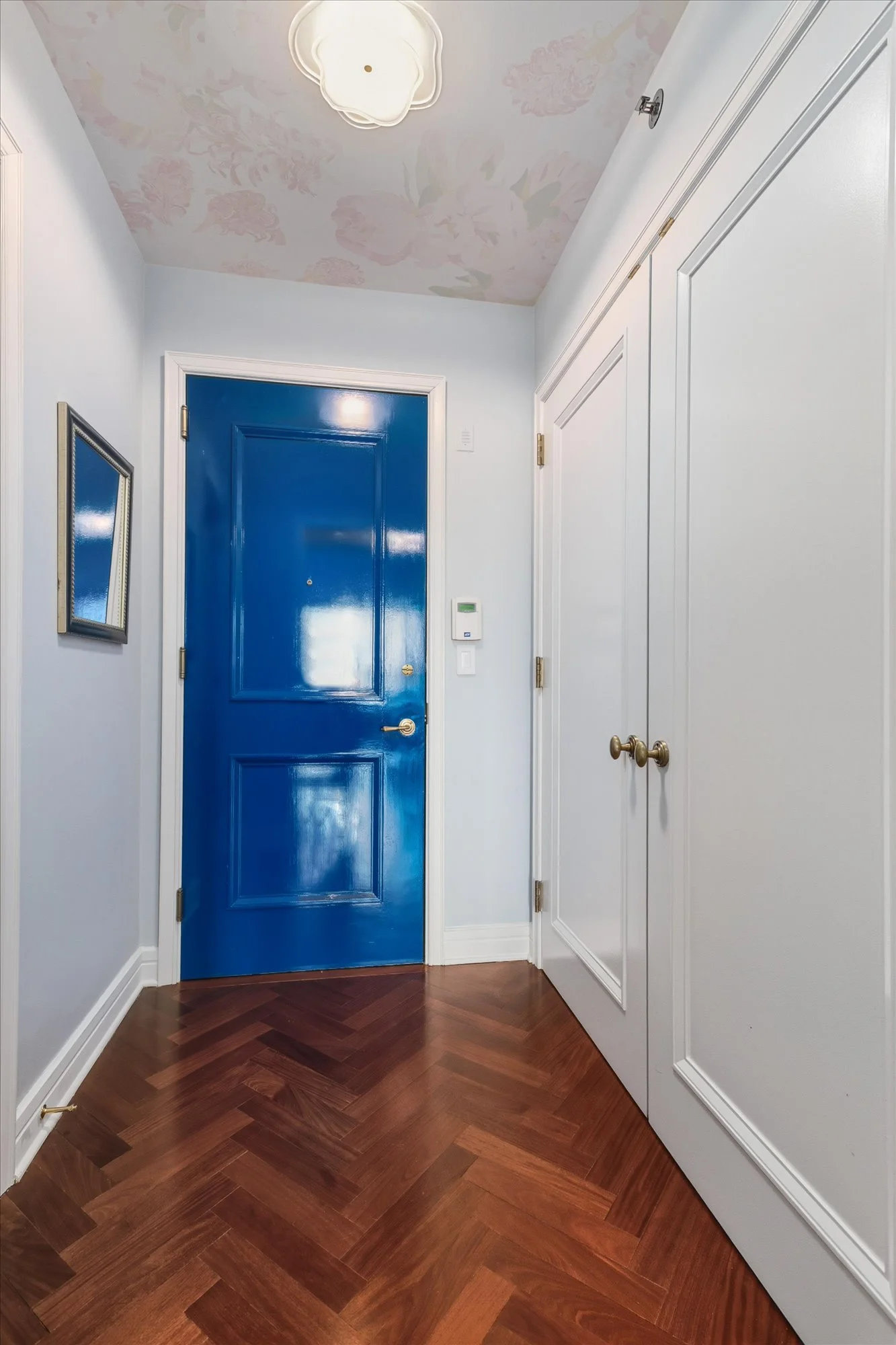 A hallway with a blue front door, white walls, a floral ceiling, and a closet with white paneled doors. The floor is made of polished hardwood in a herringbone pattern.