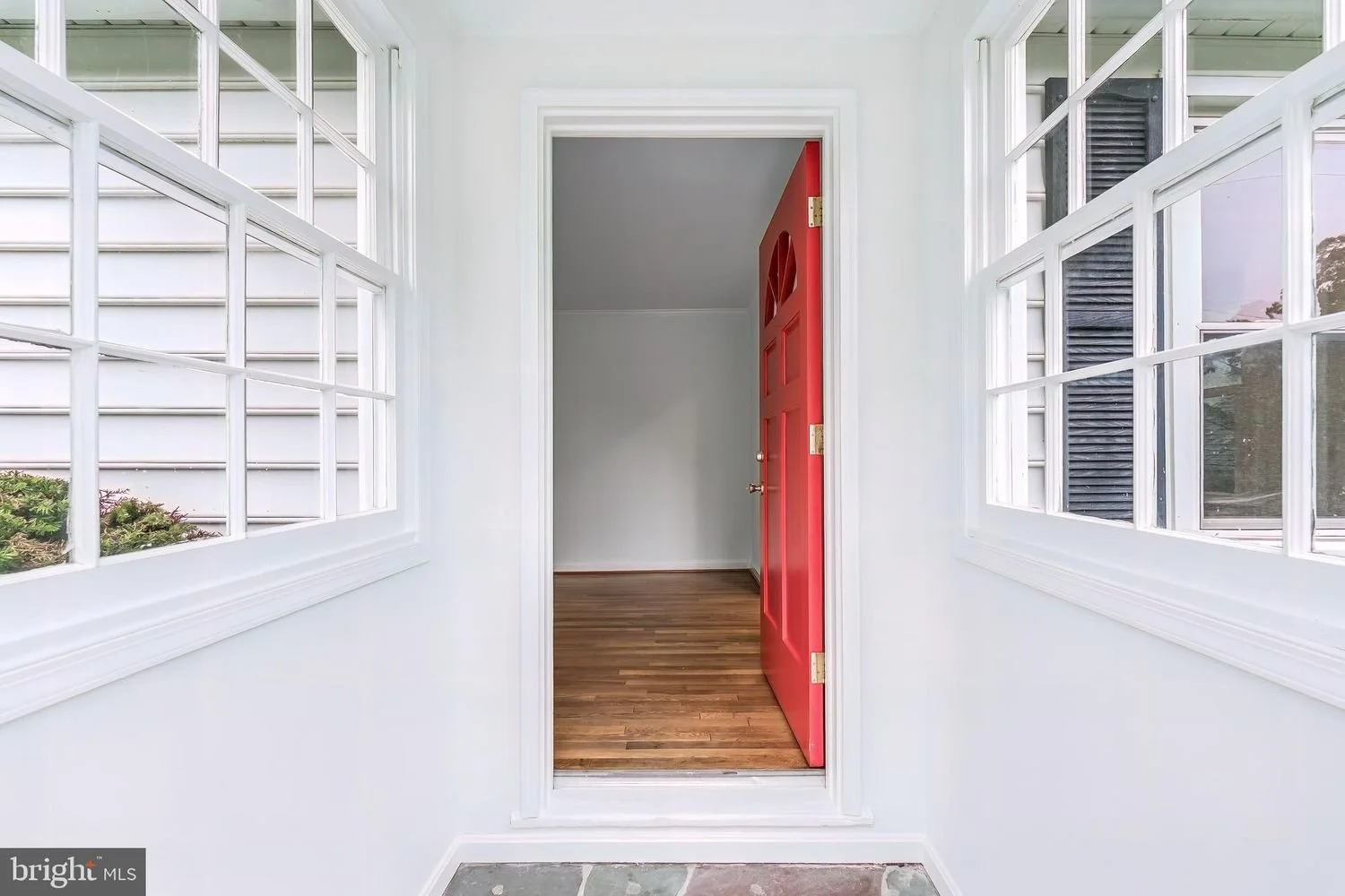 View of a front porch or entryway with a white framed screen door and windows on both sides, leading to an indoor space with hardwood flooring and a partially open red front door.