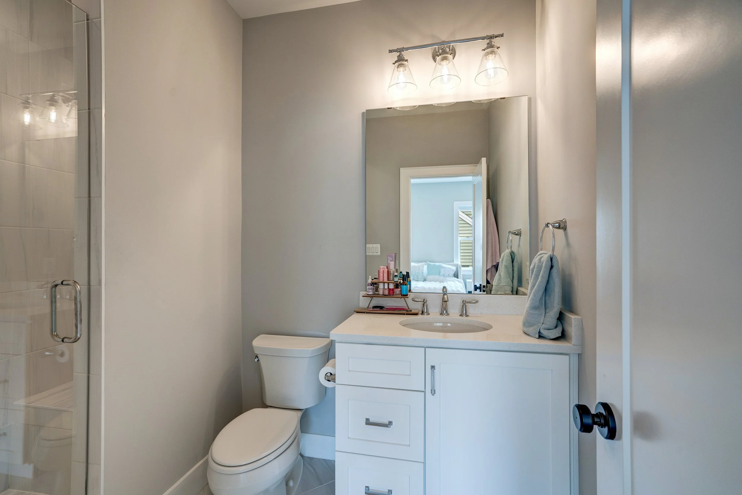 A bathroom with a white toilet, a white vanity with a sink, a mirror, and a light fixture with three glass shades. There are toiletries on the vanity and towels hanging on towel rings. A reflection of a bedroom is visible in the mirror.