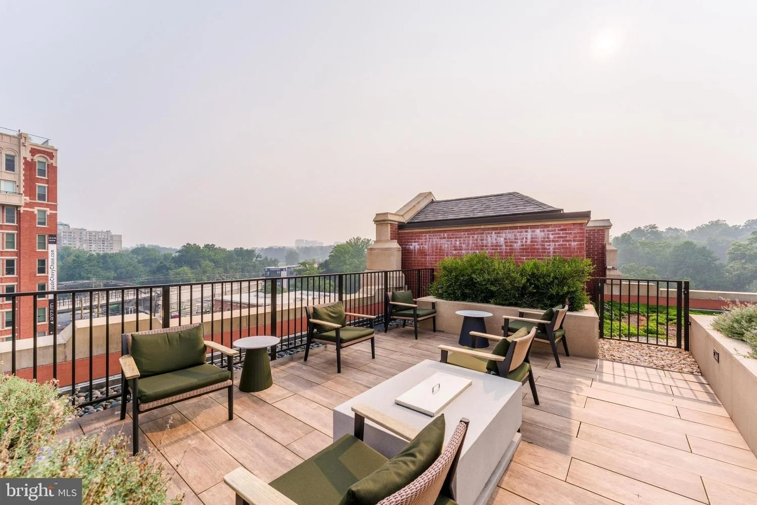 A rooftop terrace with wooden flooring, outdoor seating including chairs with green cushions, small tables, and lush green plants. City buildings and greenery are visible in the background under a clear sky.