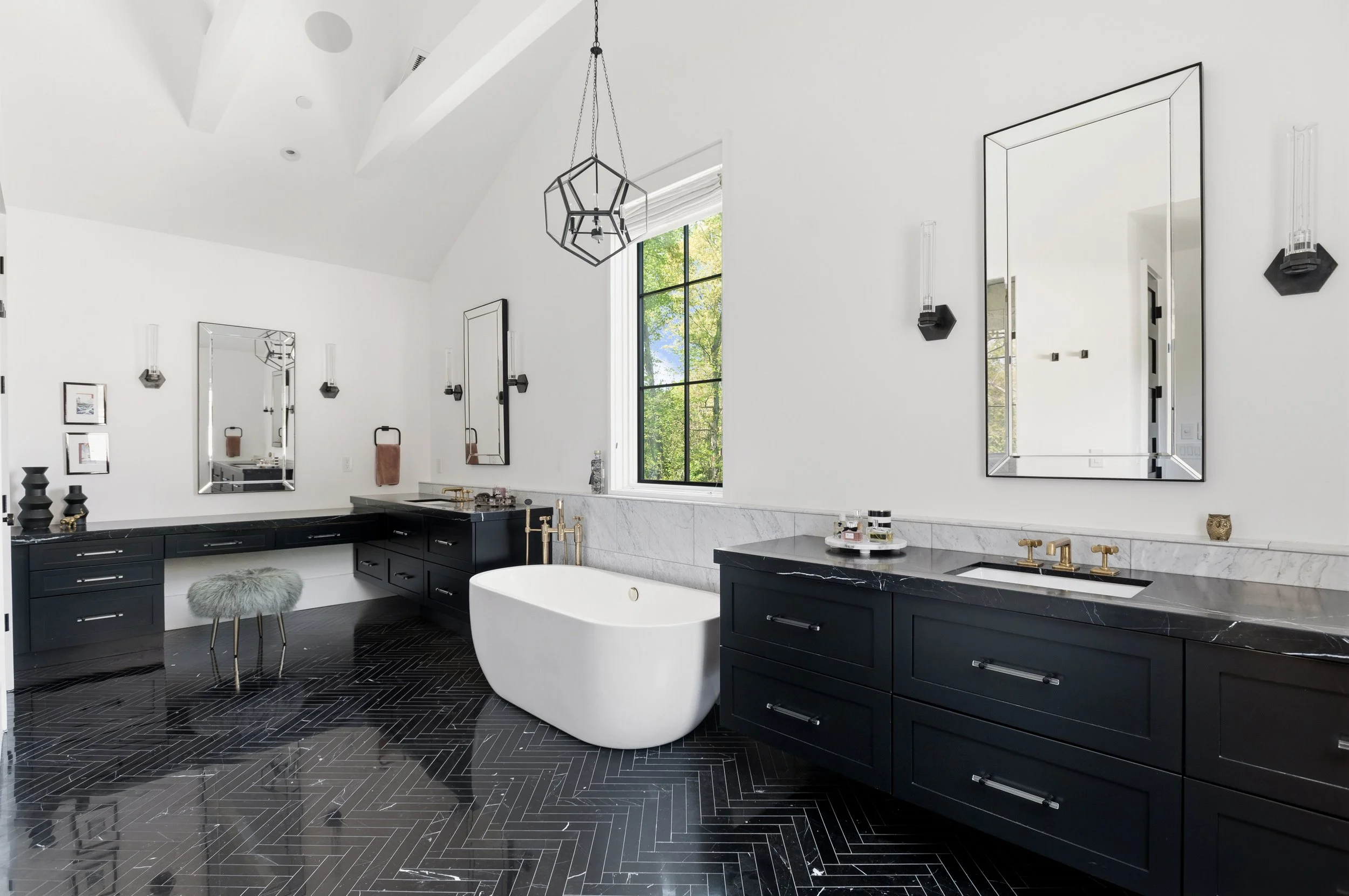 Elegant modern bathroom with black cabinetry, marble countertops, a free-standing bathtub, large mirrors, and black tile flooring.