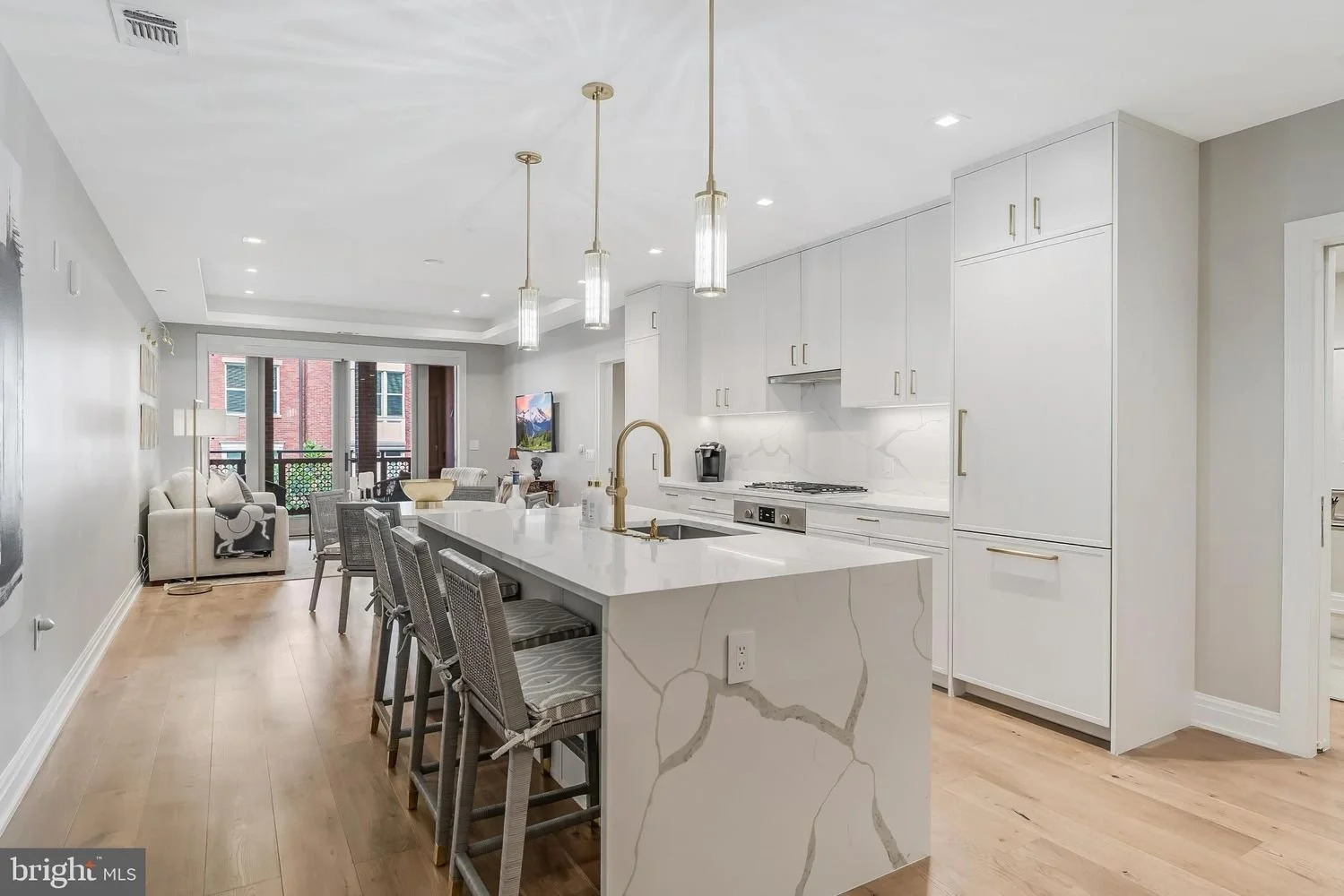 Modern open-concept kitchen with white cabinetry, marble countertops, gold fixtures, and a large island with barstools, overlooking the living area with a sofa and sliding glass doors.