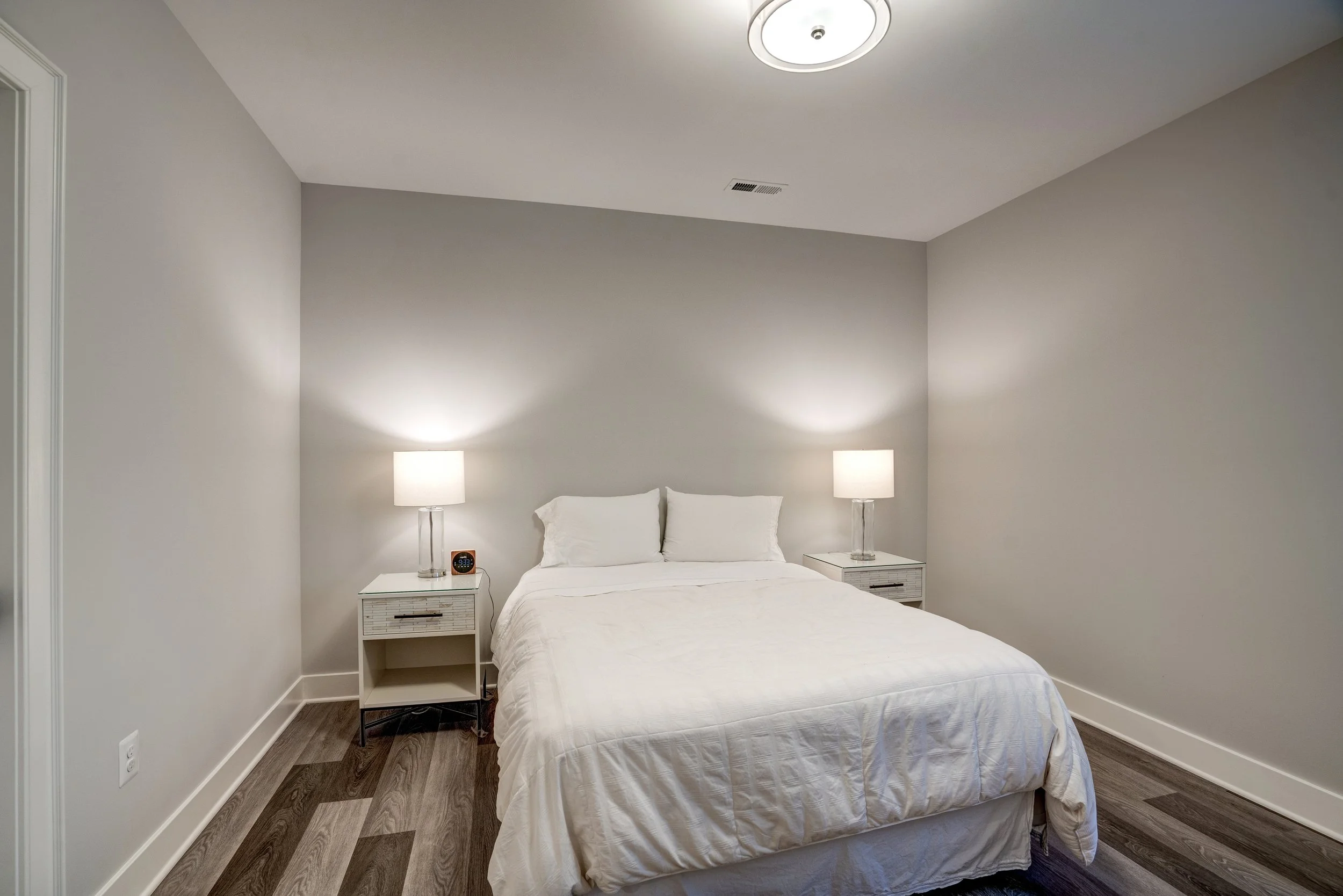 A bedroom with a bed, white linens, two white pillows, two matching nightstands with lamps, a digital alarm clock, and a ceiling light. The walls are painted light gray, and the floor is dark wood.