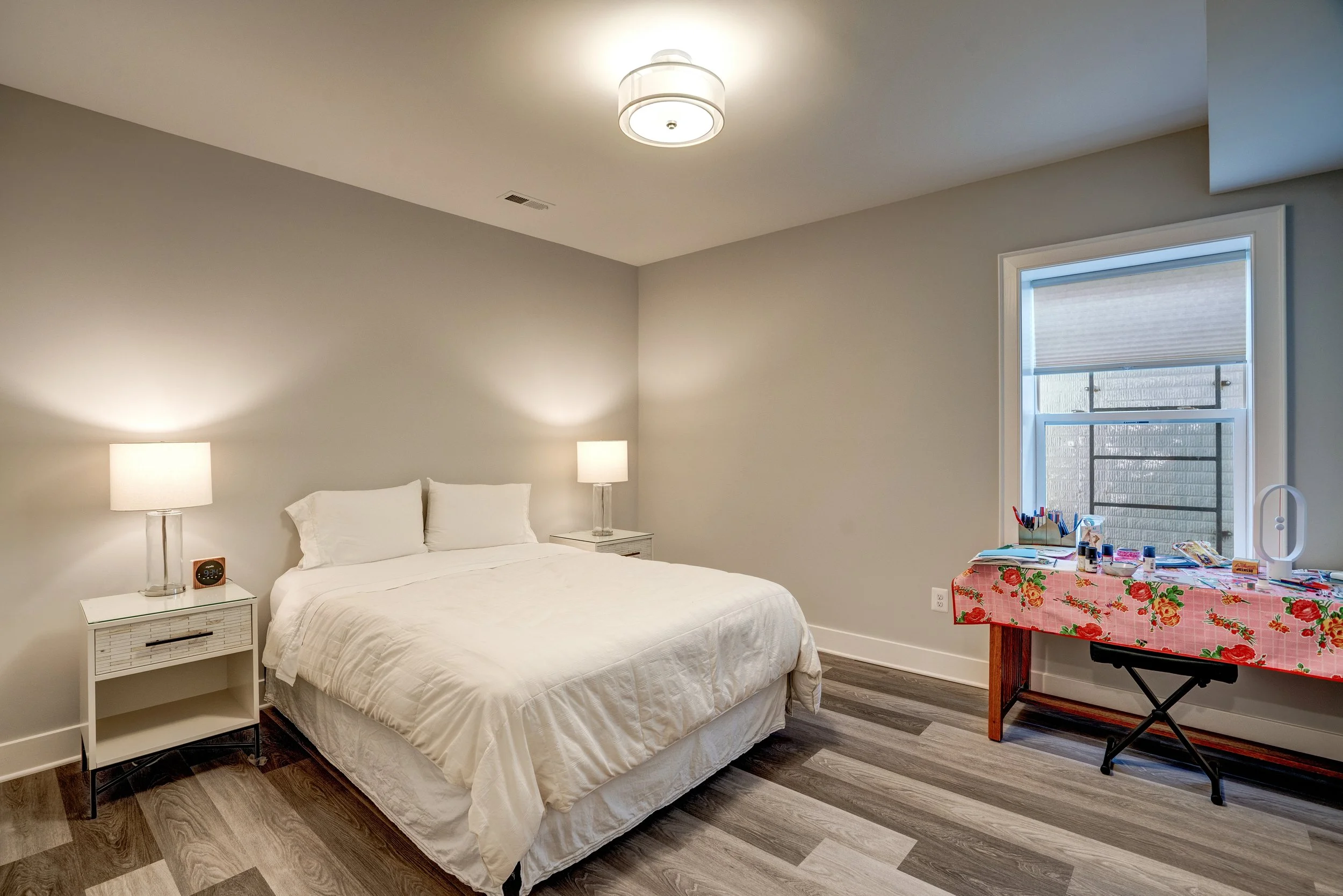 A bedroom with a white bed, two white nightstands with lamps, and a window with a roller shade. There is a table with a floral tablecloth, art supplies, and a white Dyson fan.