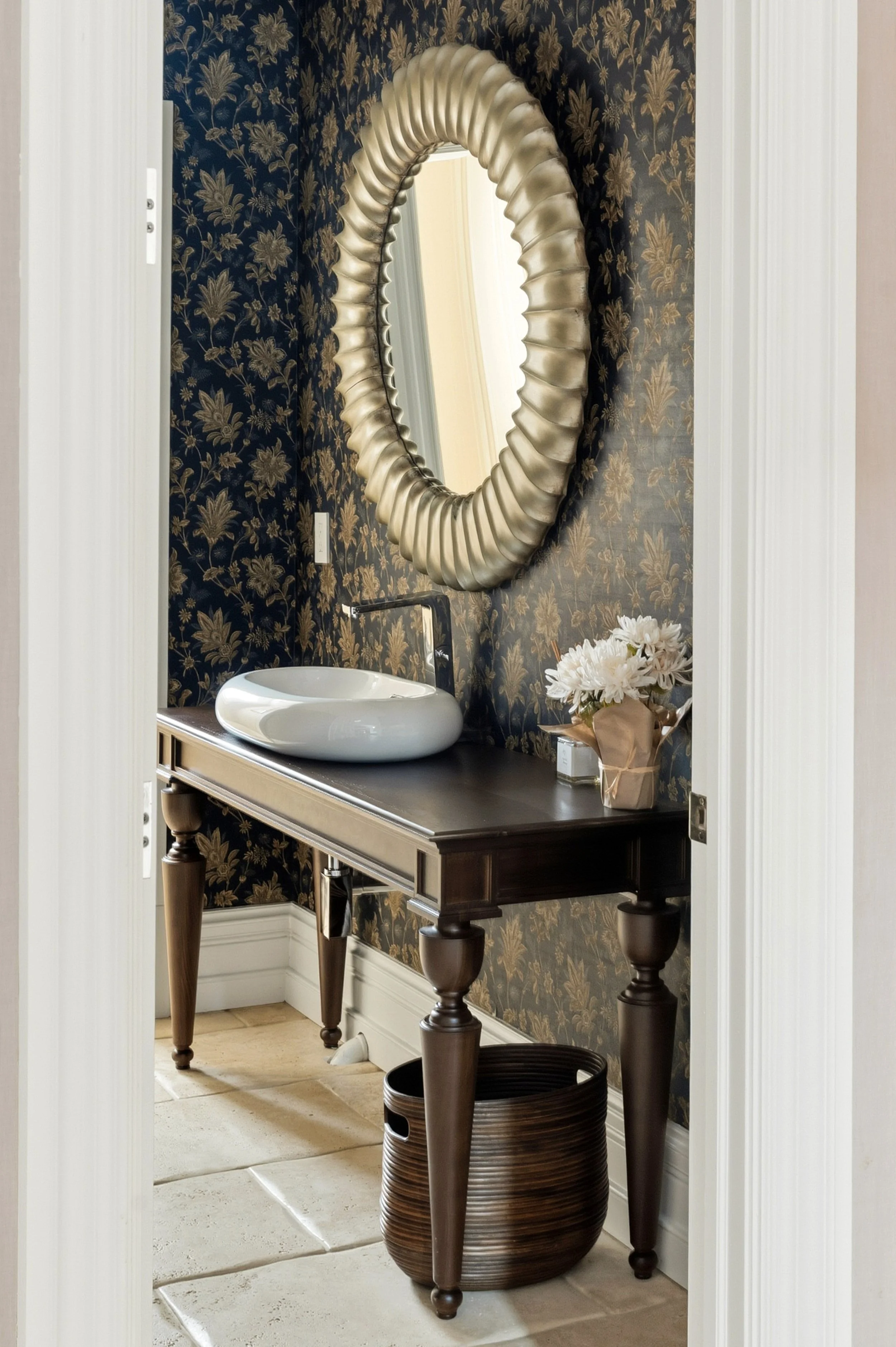 Decorative bathroom with floral wallpaper, a large ornate mirror, a white vessel sink on a dark wooden table, a bouquet of white flowers, and a woven basket, with tiled flooring.