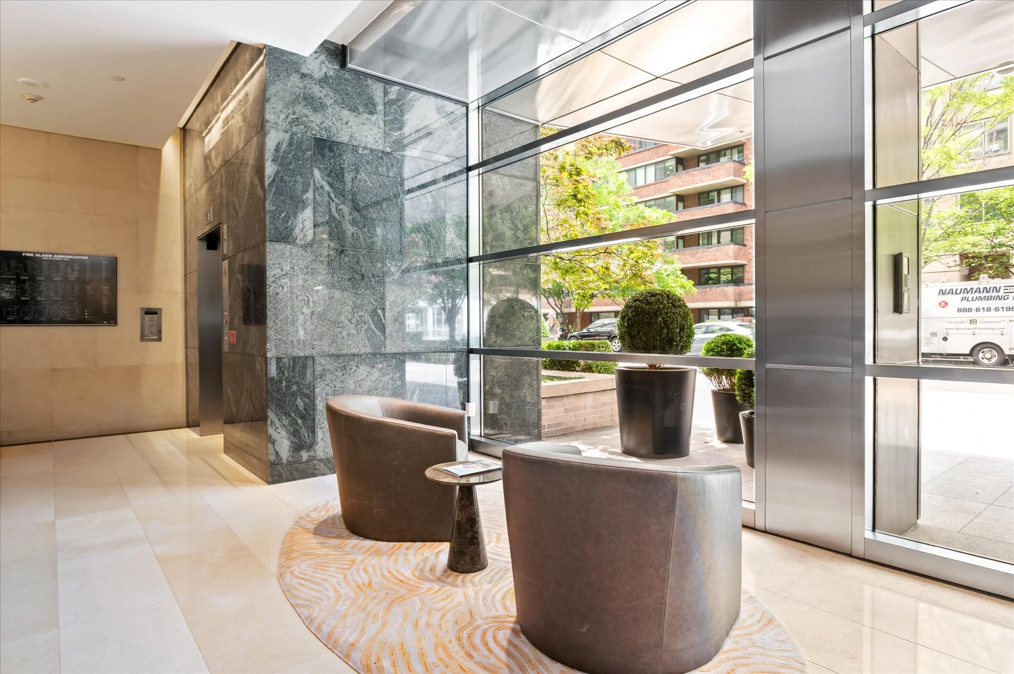 Indoor lobby area with large glass windows showing trees and buildings outside, featuring chairs, potted plants, a carpet, and an elevator.