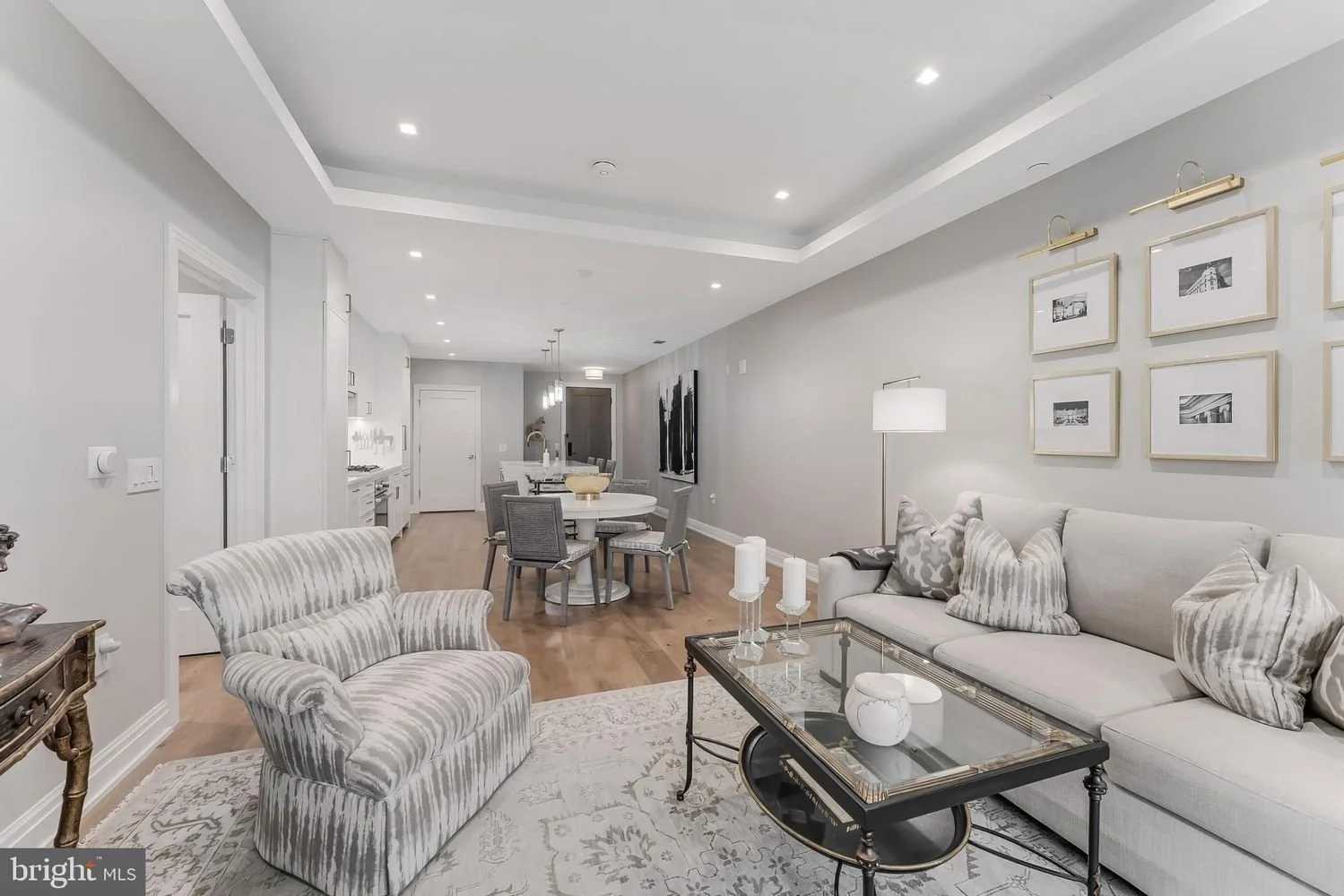 Open-concept living room and dining area with light gray walls, hardwood flooring, a striped armchair, a white sofa with patterned pillows, a glass coffee table, a standing lamp, framed black-and-white photographs, a round dining table with gray chai