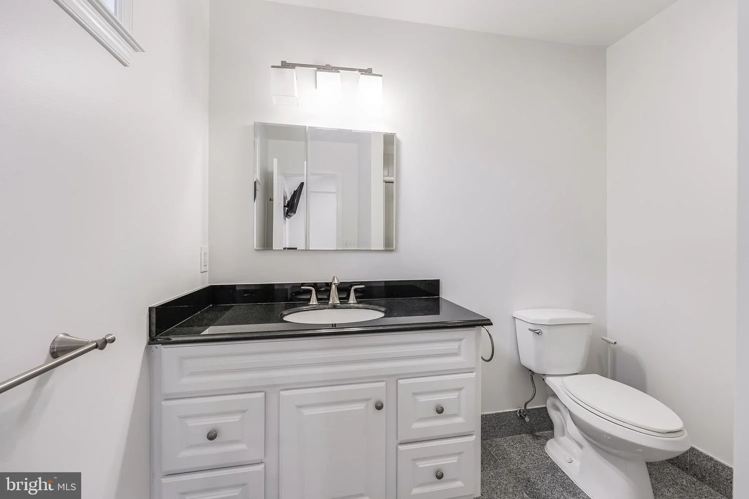 A clean and modern bathroom with white walls, a black countertop vanity with a built-in sink, a rectangular mirror above the sink, a wall-mounted light fixture, a white toilet, and a granite floor.