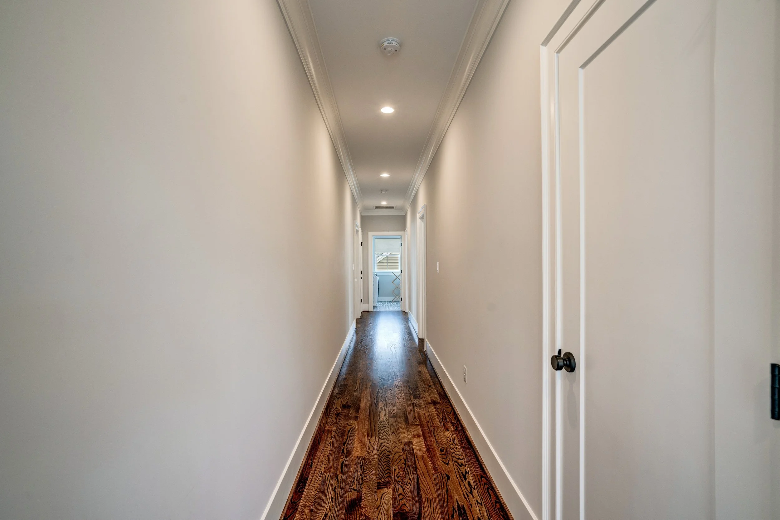 Empty hallway with wooden floor, white walls, and ceiling lights. A door is partially visible on the right, and a window can be seen at the end of the hallway.