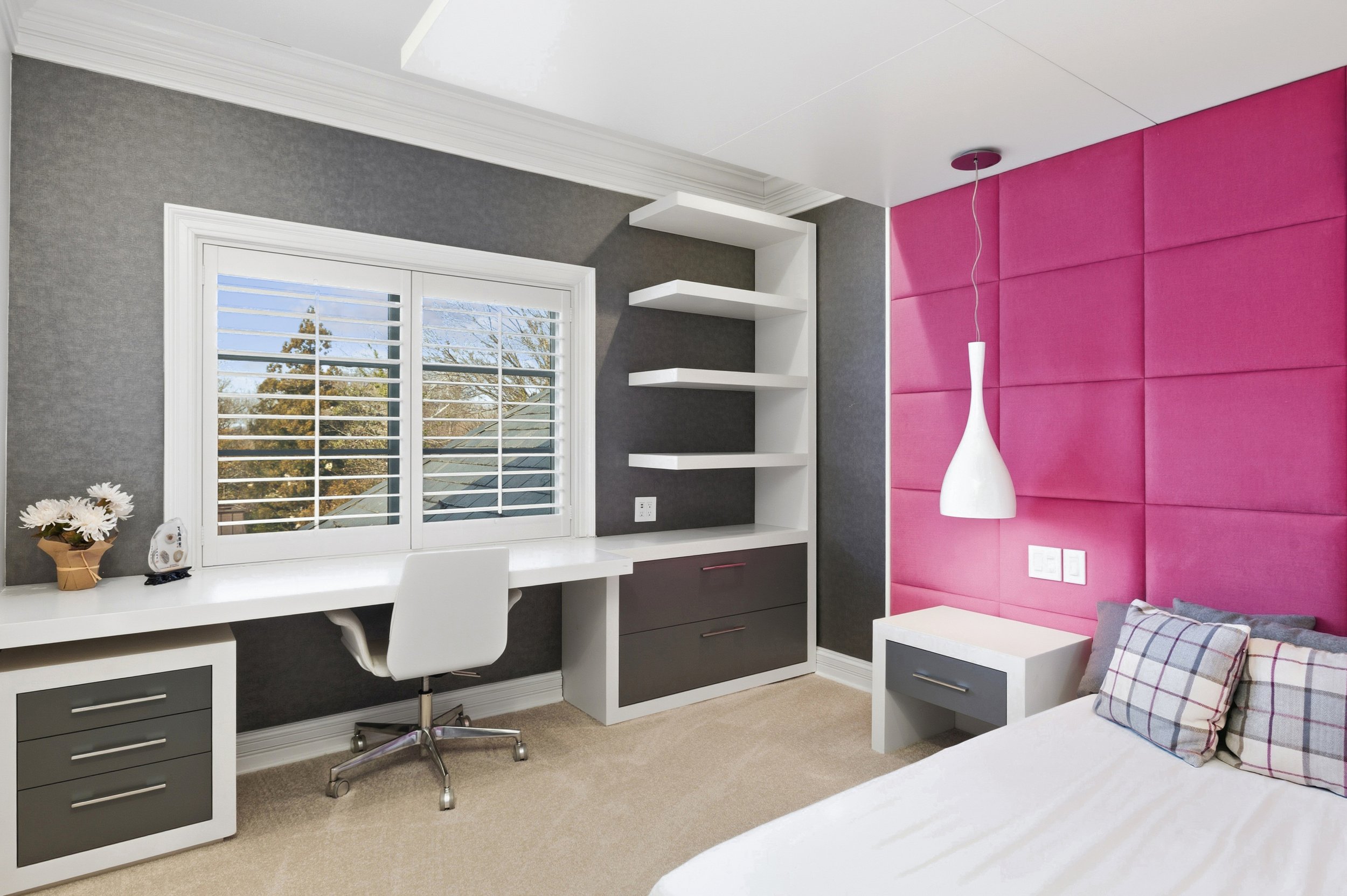 A modern bedroom with a pink padded accent wall, a white ceiling lamp, white and gray furniture, a window with white shutters, and a desk with a computer and decorative items.