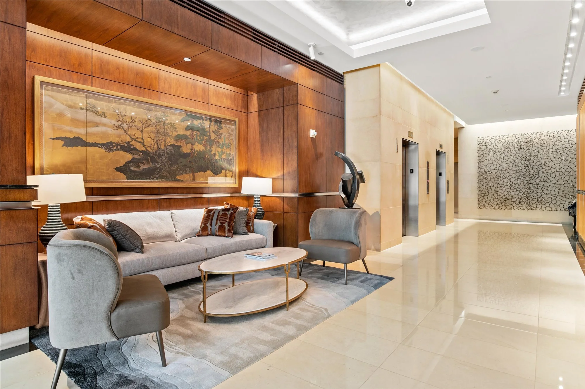 Modern hotel lobby with wooden wall panels, a large framed Asian-style painting, a beige sofa with decorative pillows, two light-colored armchairs, a small round coffee table, elevator doors, and a textured wall art piece.