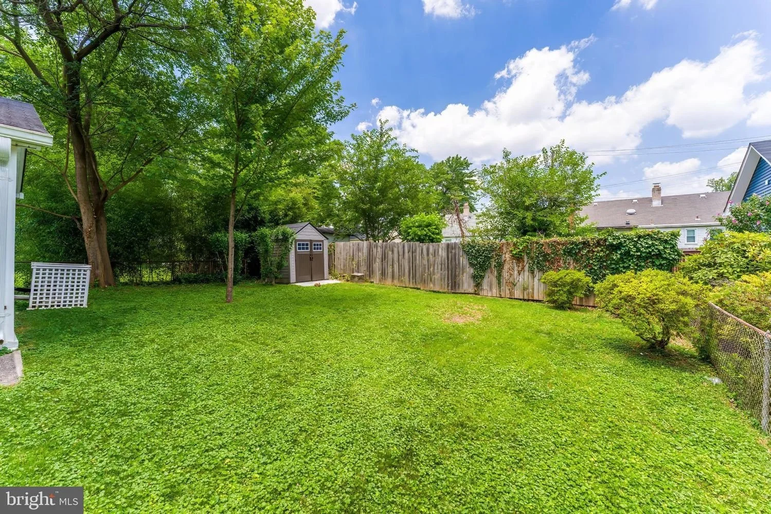 A spacious backyard with bright green grass, trees, a wooden shed, a wooden fence, and shrubbery under a partly cloudy sky.