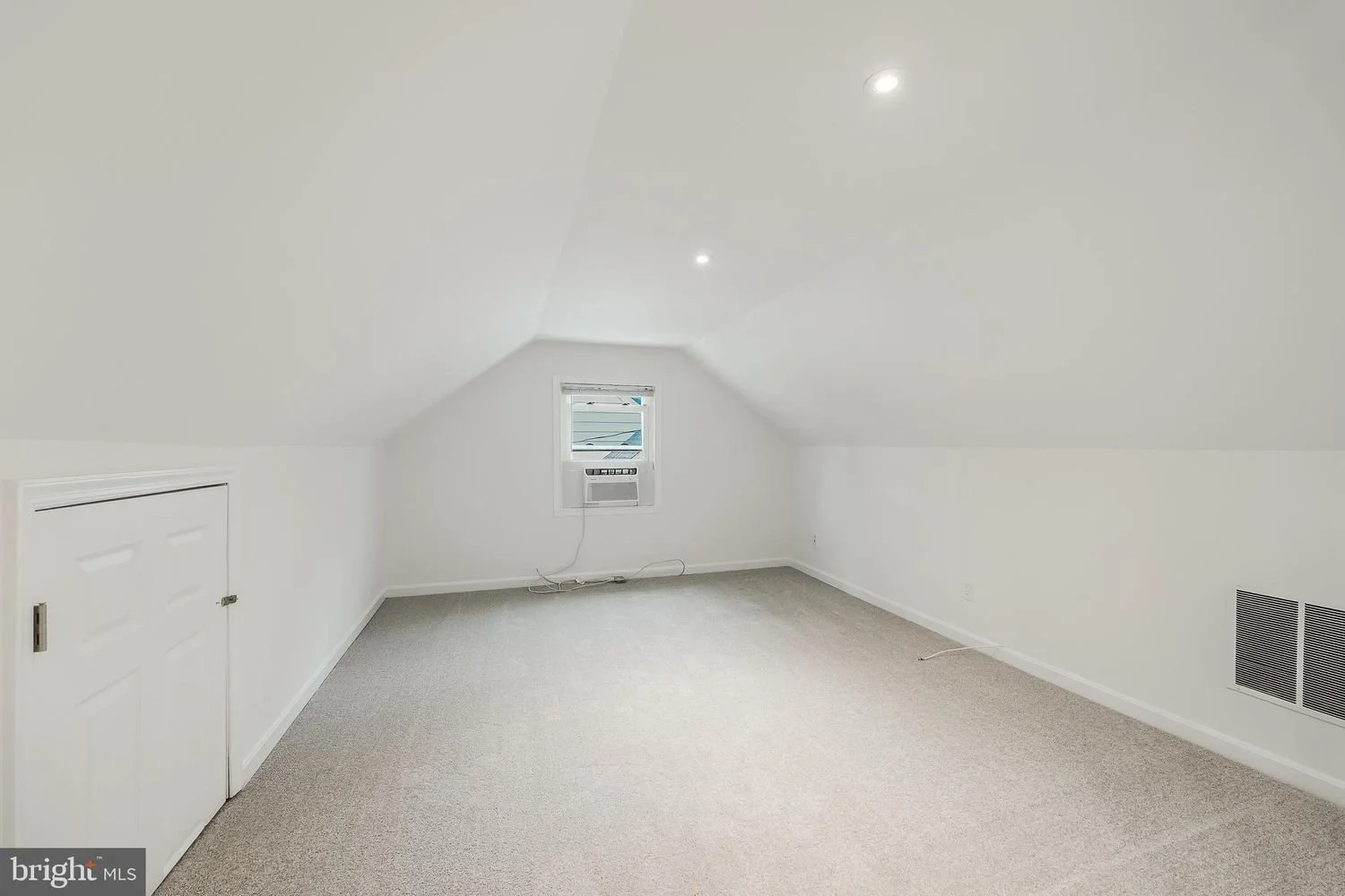 Empty attic room with sloped ceiling, white walls, beige carpet, small window with window air conditioning unit, and ventilation grille on the wall.