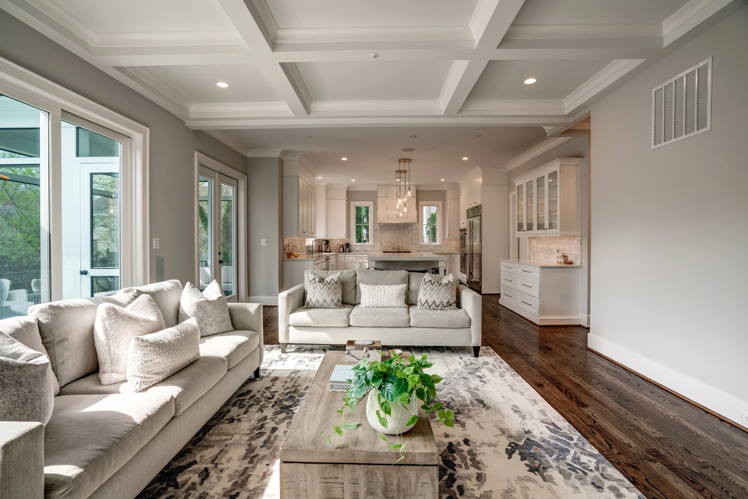 Living room with a beige sofa, a coffee table with a plant, and large windows showing an outdoor patio.