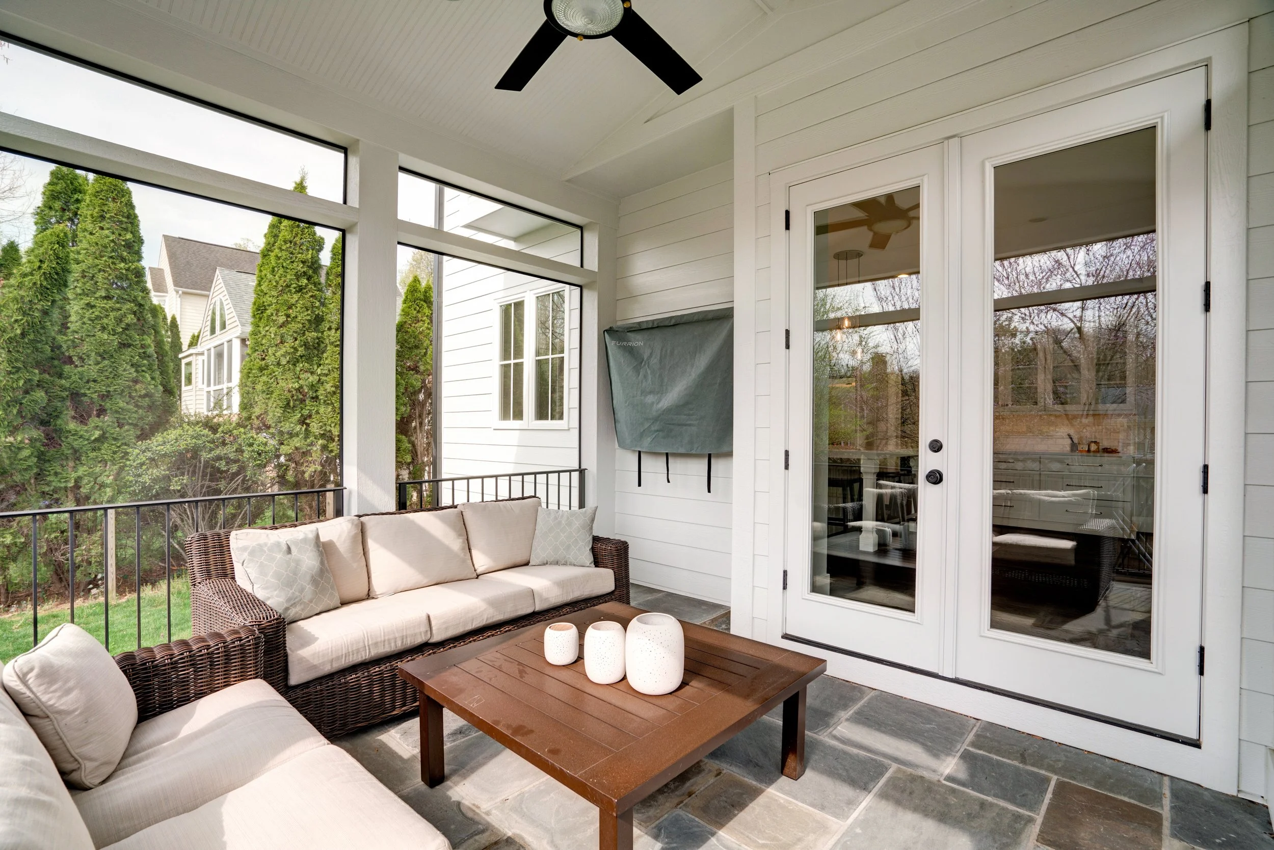 Covered porch area with beige outdoor sofa, dark brown wicker chairs, and a wooden coffee table with white decorative vases. White siding walls and glass double doors lead inside. Ceiling fan with black blades and a glass light fixture hang from the 