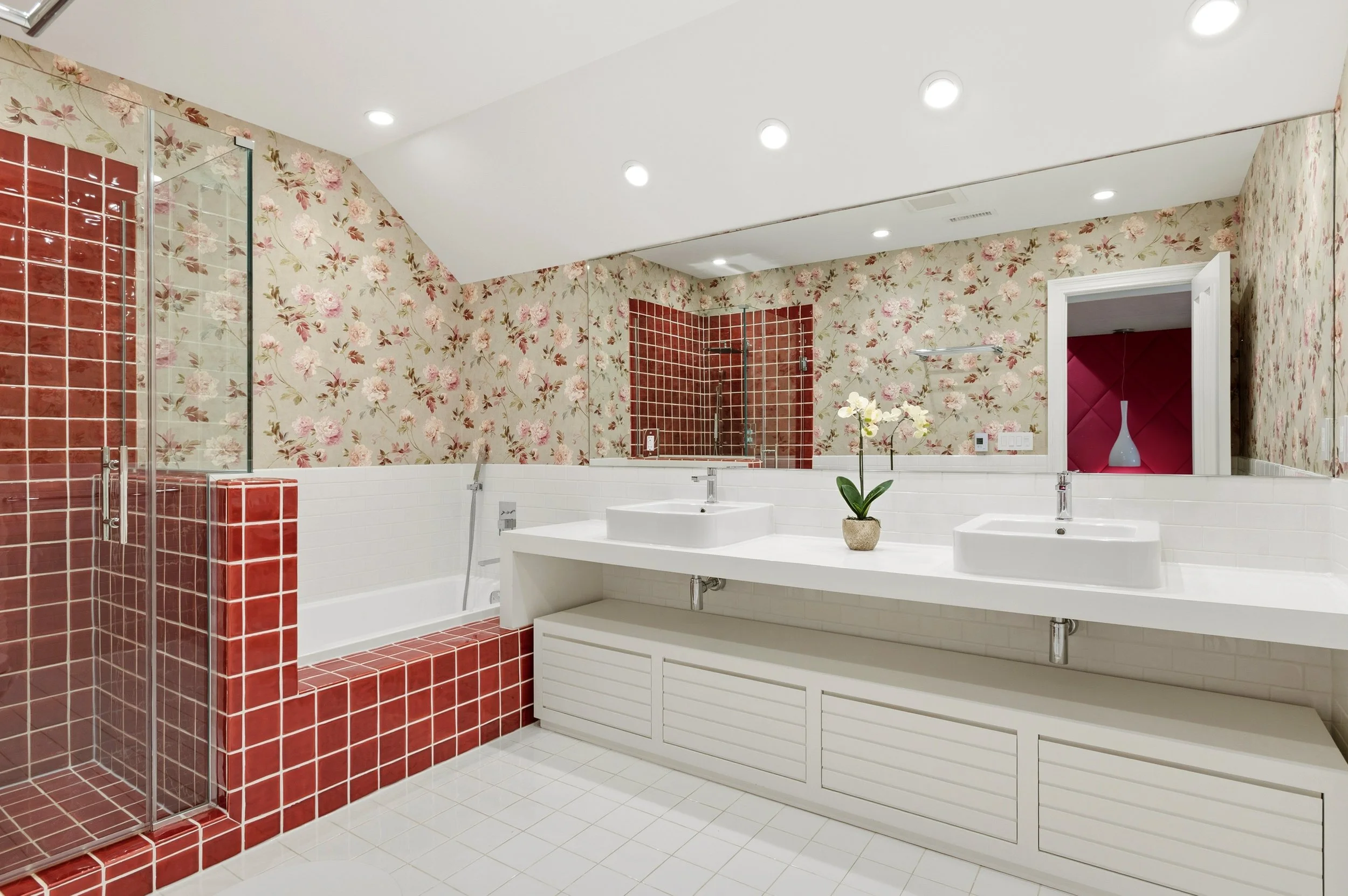 A bathroom featuring floral wallpaper, red tiled shower with glass enclosure, white double sinks with a plant in the center, and a large mirror with recessed ceiling lights.