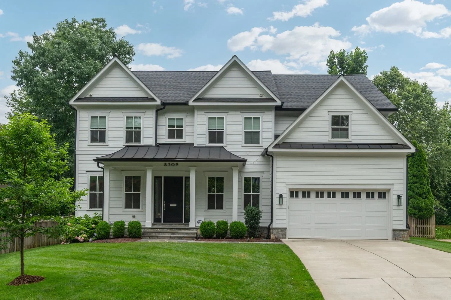 A large white two-story house with black roof shingles and a two-car garage, surrounded by a green lawn and trees under a partly cloudy sky.