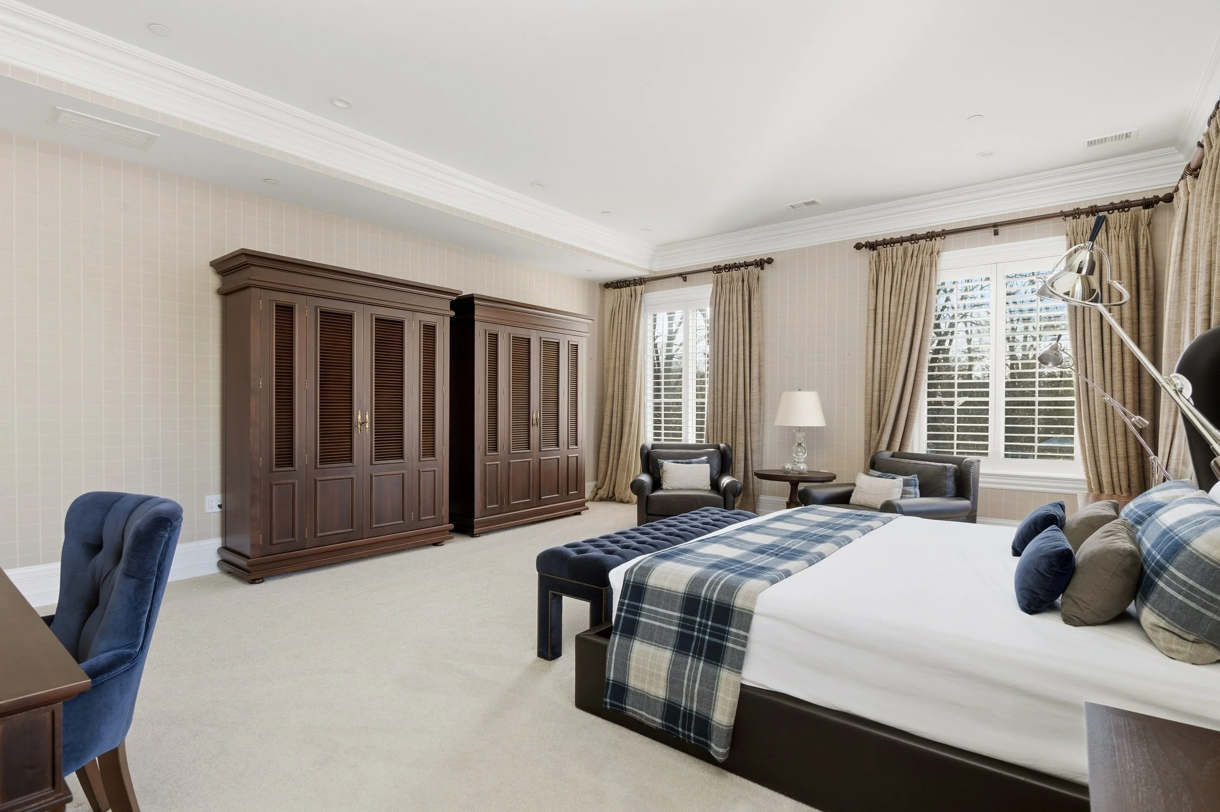 A well-lit bedroom with large windows, beige curtains, a white bed with plaid bedding, dark wood furniture, and a seating area with armchairs and a side table.