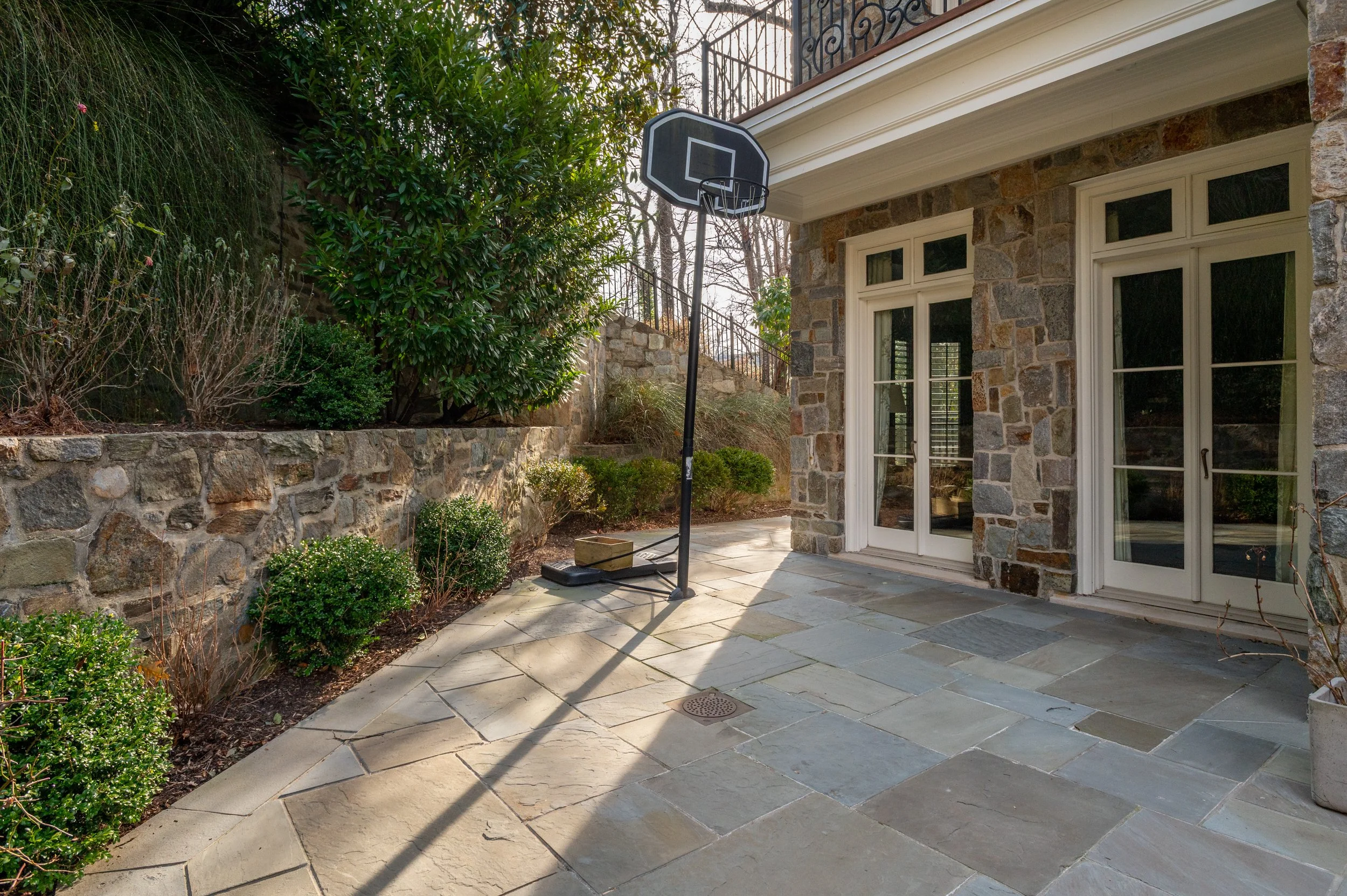 A backyard patio with a basketball hoop, stone walls, and a sliding glass door on a stone house, surrounded by greenery and bushes.