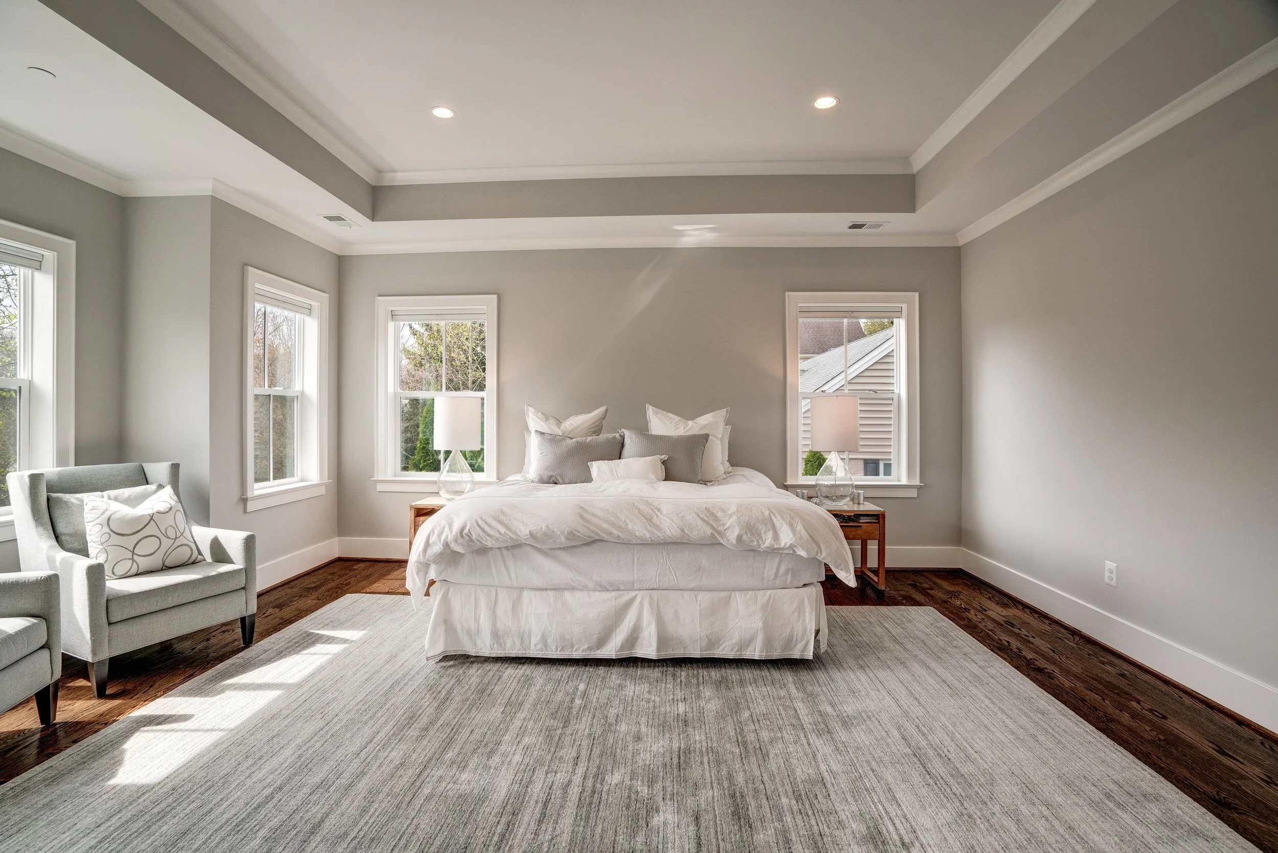 A bedroom with a white bed, gray throw pillows, and bedside tables with lamps. There are three windows with white frames letting in natural light, and a gray armchair with a patterned pillow near the bed.