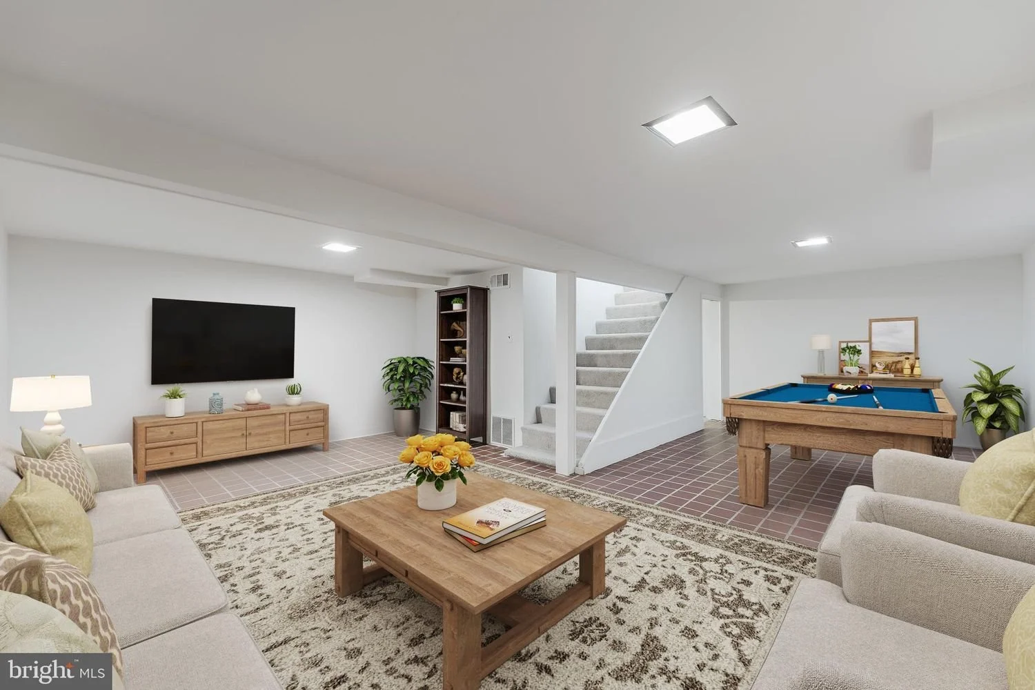 Basement living room with beige sofas, a wooden coffee table with books and a yellow flower arrangement, a flat-screen TV on a wooden TV stand, a pool table with cues, potted plants, and a staircase.
