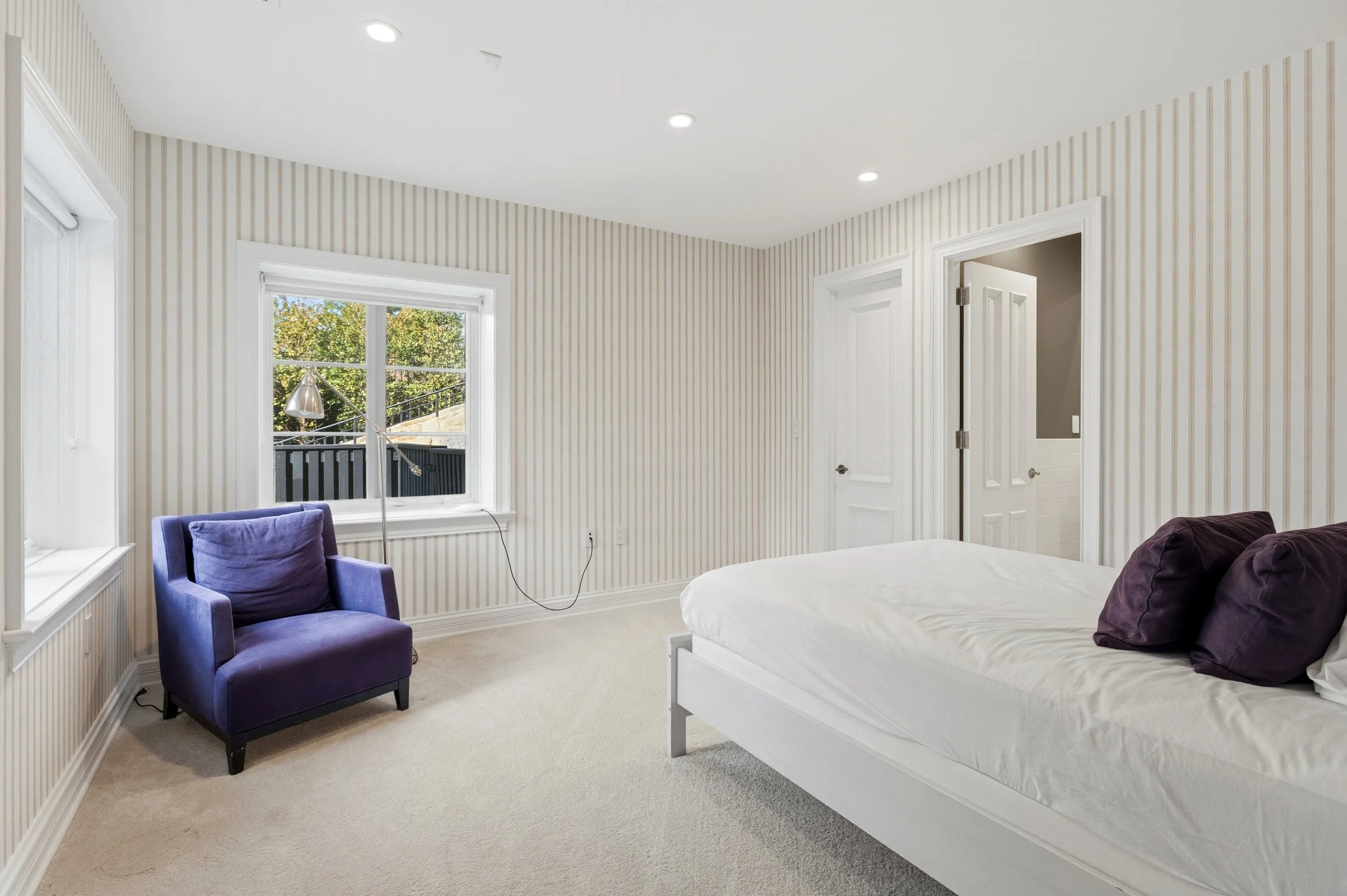 A bedroom with beige striped wallpaper, a white bed with dark pillows, purple armchair, window with view of trees, and a white door.
