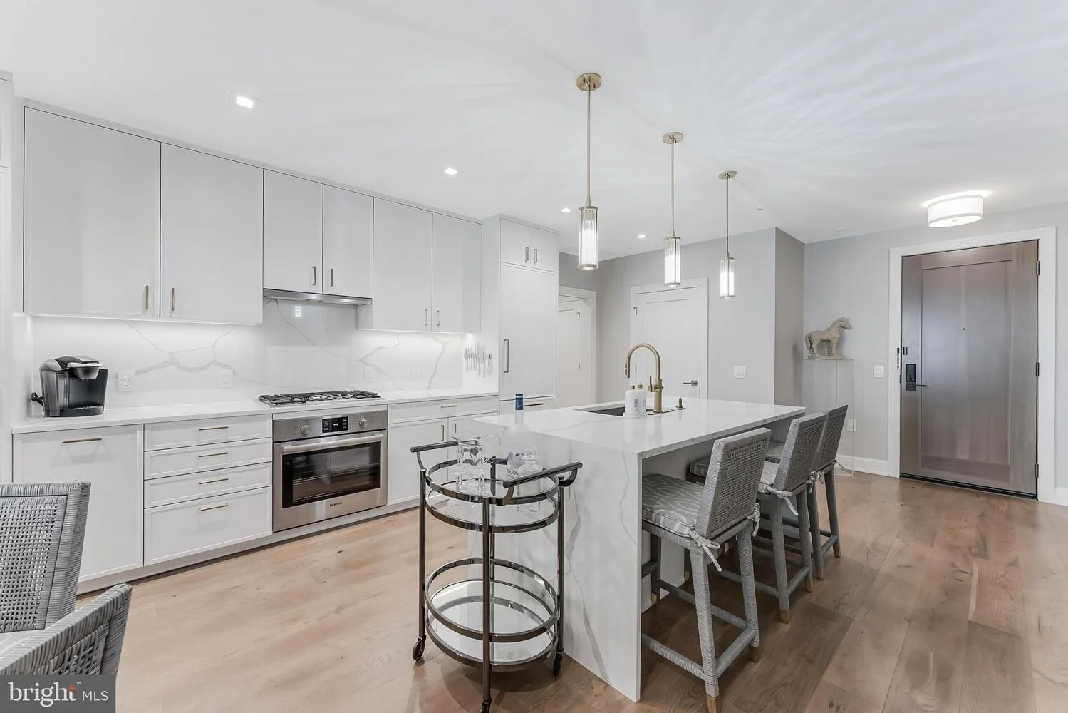 Modern kitchen with white cabinets, marble countertops, and a kitchen island with seating. There is a built-in oven, gas stove, and coffee maker, with hardwood floors and pendant lighting.