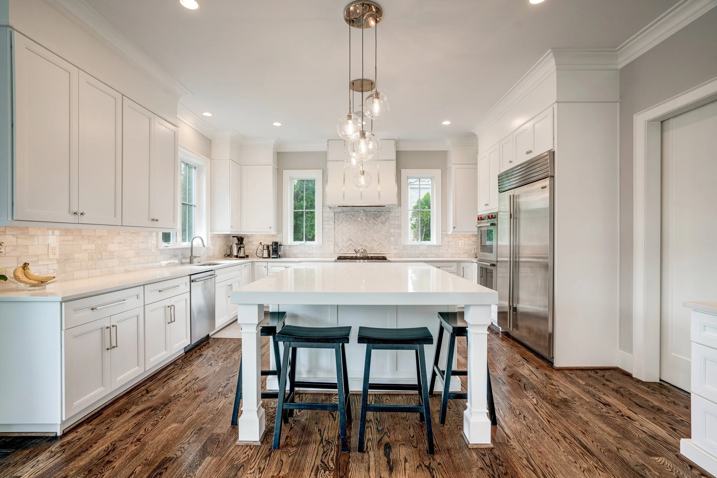 Modern white kitchen with wood flooring, central island with four black stools, white cabinets, stainless steel appliances, a window with greenery outside, and decorative pendant lights.