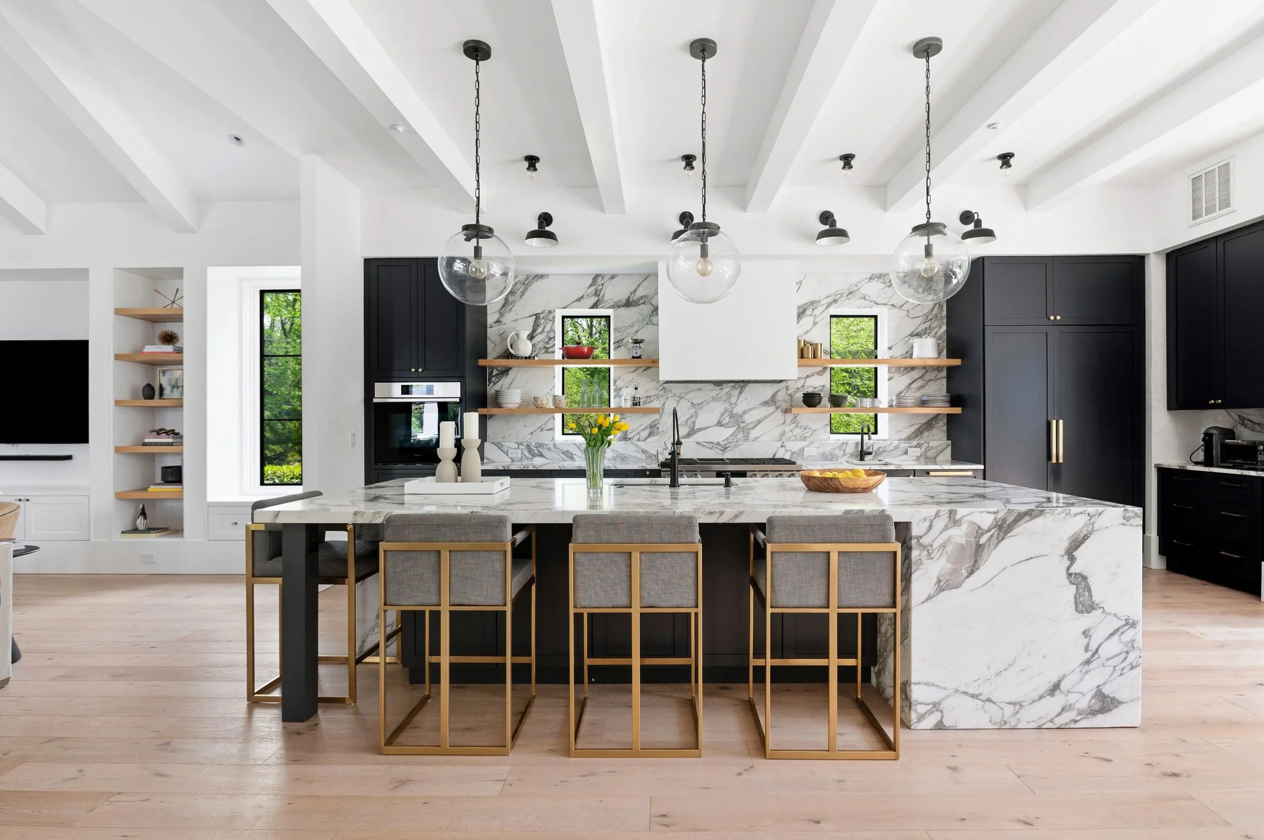 Modern kitchen with black cabinets, marble backsplash and island, wooden shelves with decor, pendant lighting, and a view of greenery outside through windows.