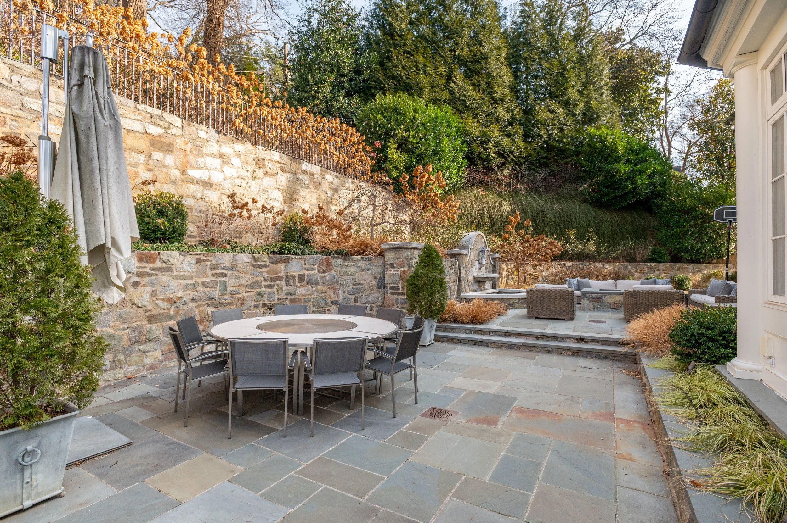 A spacious backyard patio with stone flooring, outdoor furniture including a round table with black chairs, a seating area with wicker couches and cushions, plants, and a stone wall with dry landscape and green trees in the background.