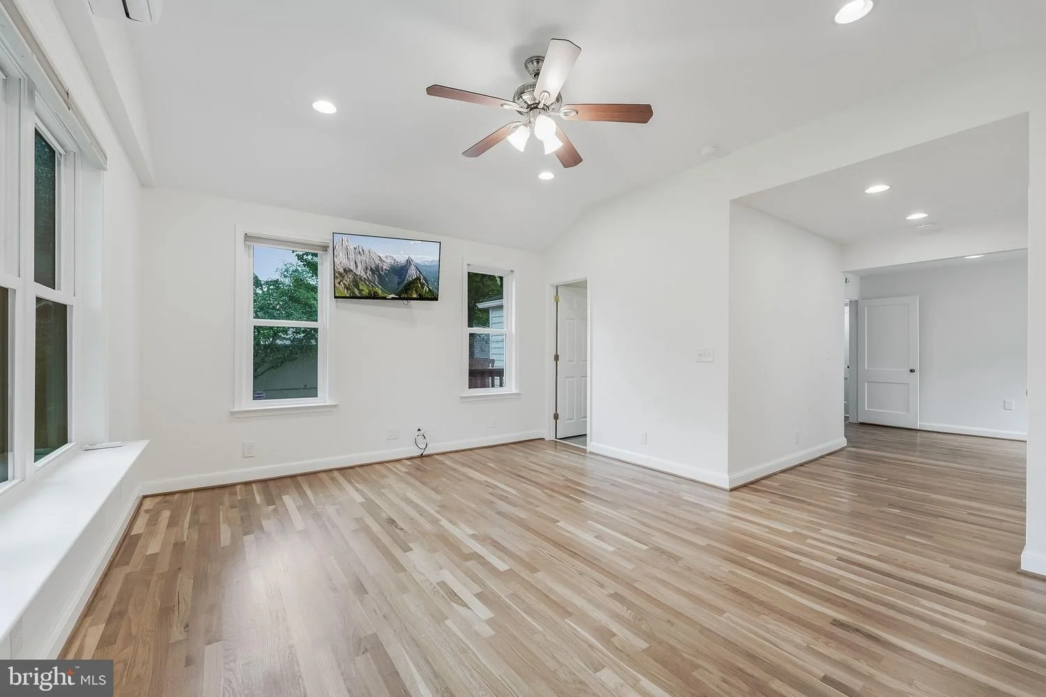 Empty living room with white walls, hardwood floors, a ceiling fan, and a wall-mounted TV.