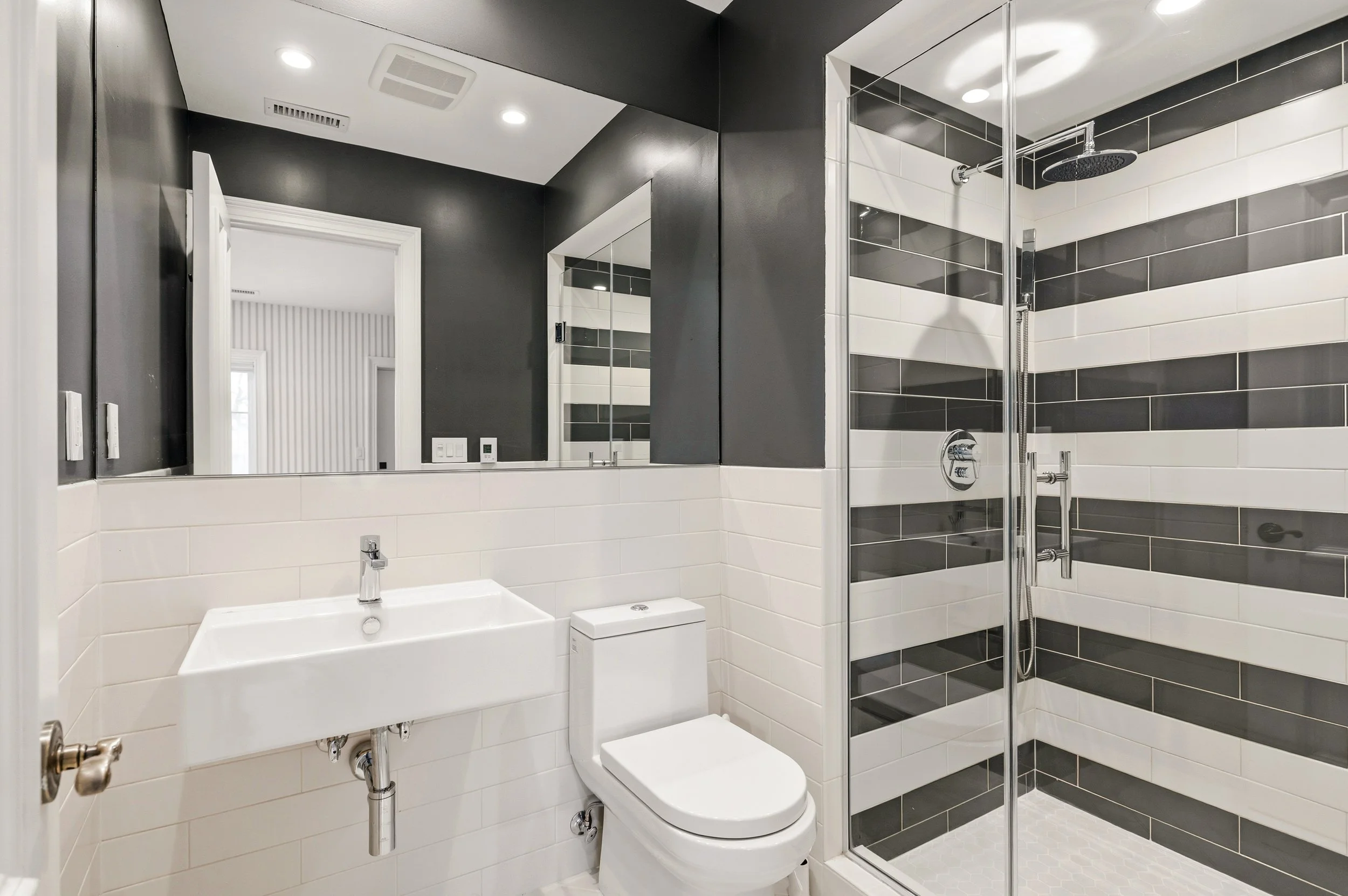 Modern bathroom with black and white tile walls, a walk-in shower with a rainfall showerhead, a white vanity with a rectangular sink, a mirror above the sink, and LED ceiling lights.
