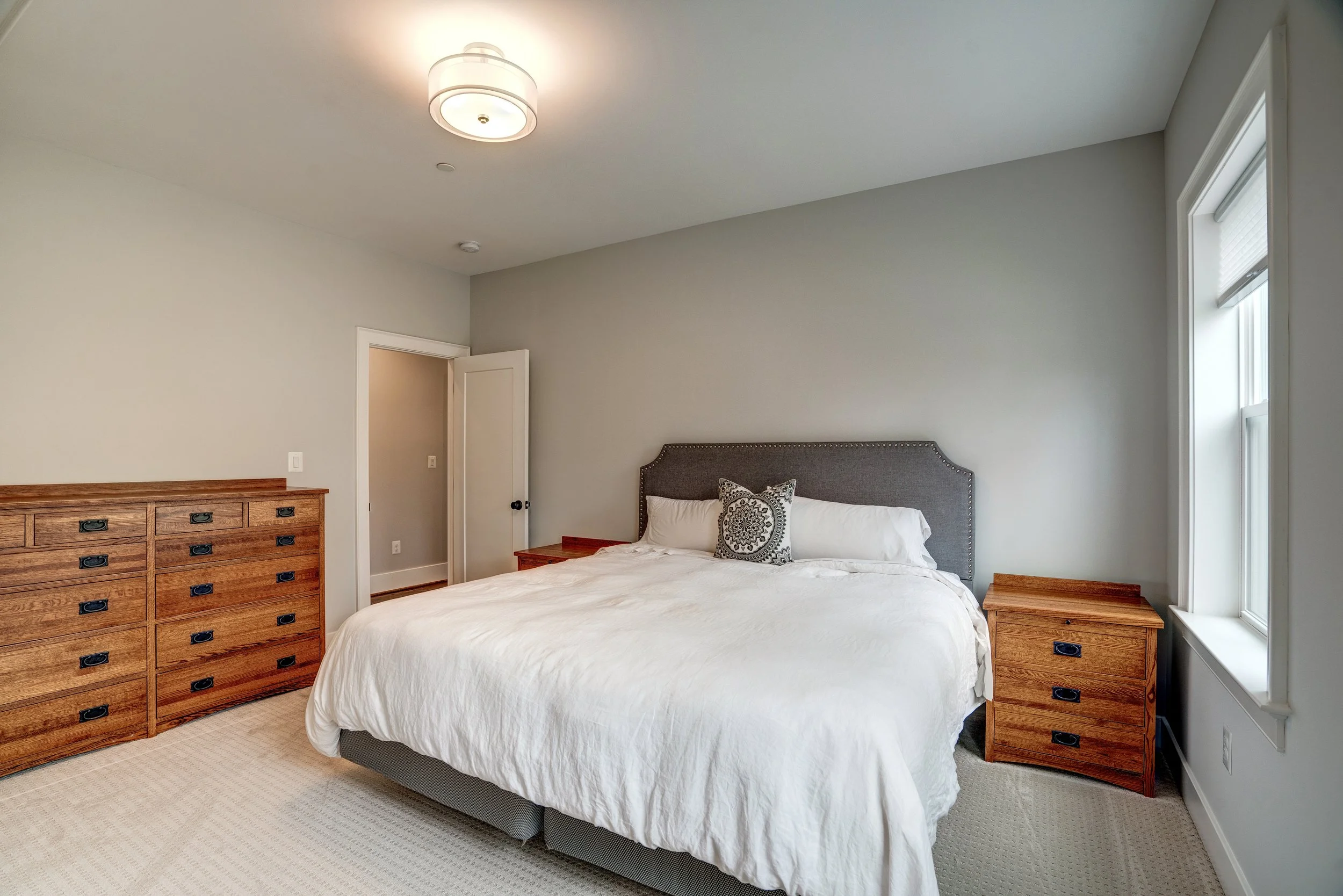 A bedroom with a bed that has a gray upholstered headboard, white bedding, and a decorative pillow. There are two wooden nightstands on either side of the bed, with a dresser on the left side. The room has a window on the right and an open door on th