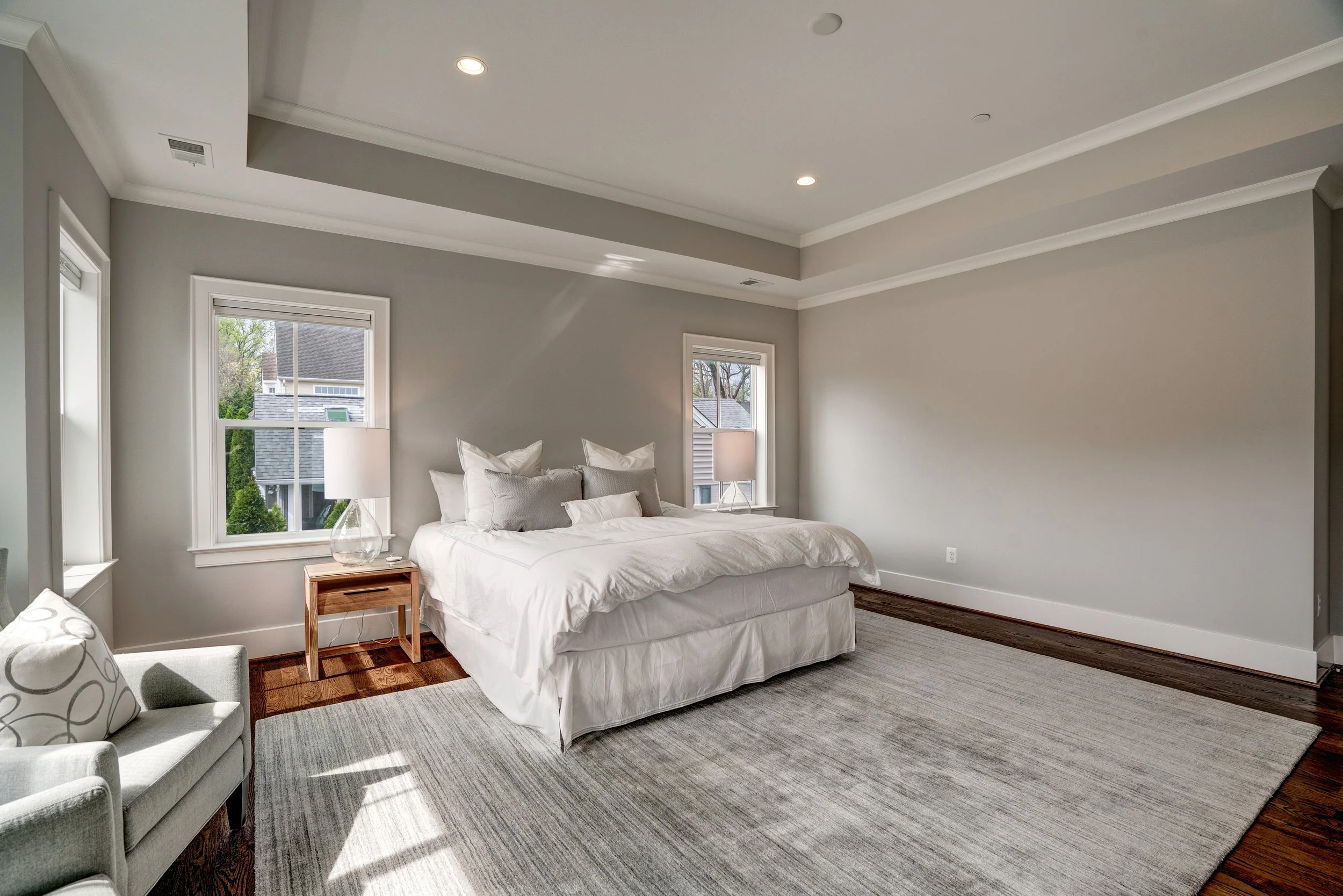 Bright bedroom with white bed, pillows, and bedside table with lamp, large windows showing neighborhood, white walls, hardwood floors, and a cozy seating area with a sofa and patterned pillows.