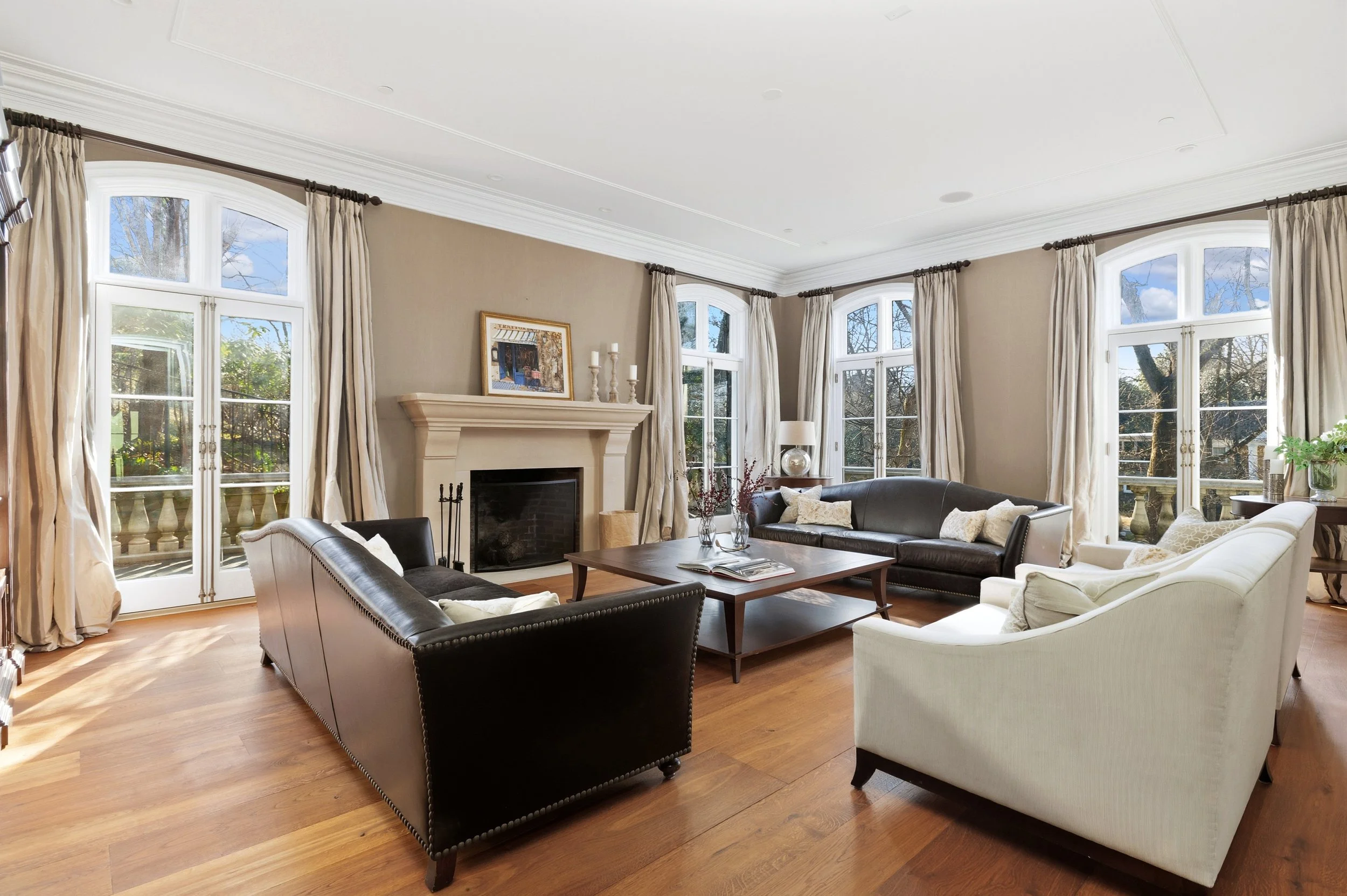A spacious living room with large windows and cream-colored curtains. There is a fireplace with a painting above it, surrounded by sofas and armchairs. A wooden coffee table with books and vases is in the center, and a small side table with a lamp is