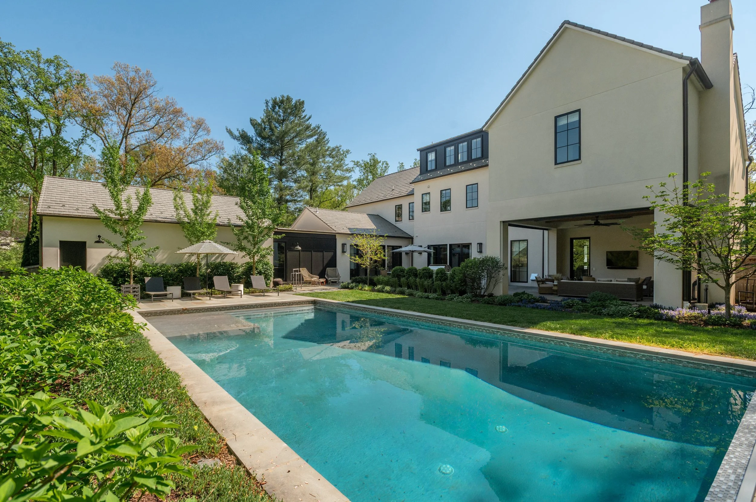 Modern house with a backyard pool, surrounded by green trees, with patio furniture and umbrellas, under a clear blue sky.