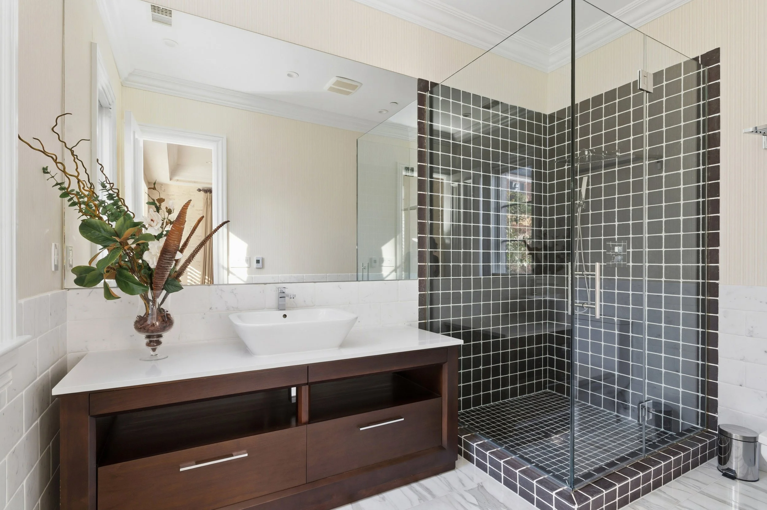 Modern bathroom with a dark wood vanity, white sink, floral arrangement, and a glass-enclosed shower with black square tiles.