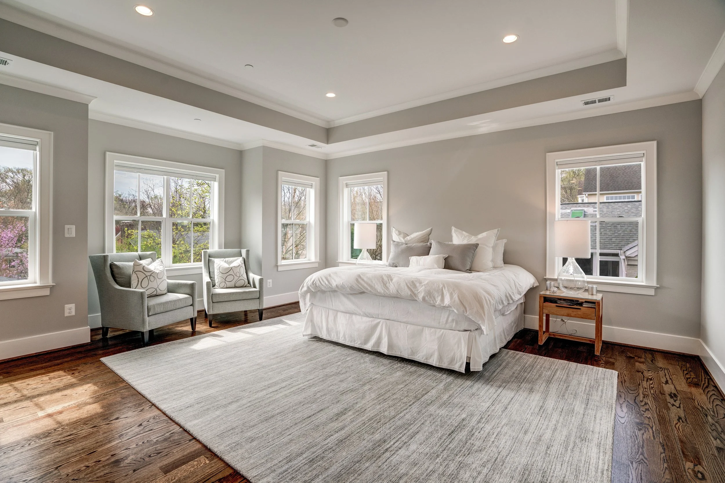 Bright bedroom with large windows, white bedding, and a wooden nightstand with a lamp. Two armchairs with pillows are near the windows, and there is a light-colored area rug on dark hardwood floors.