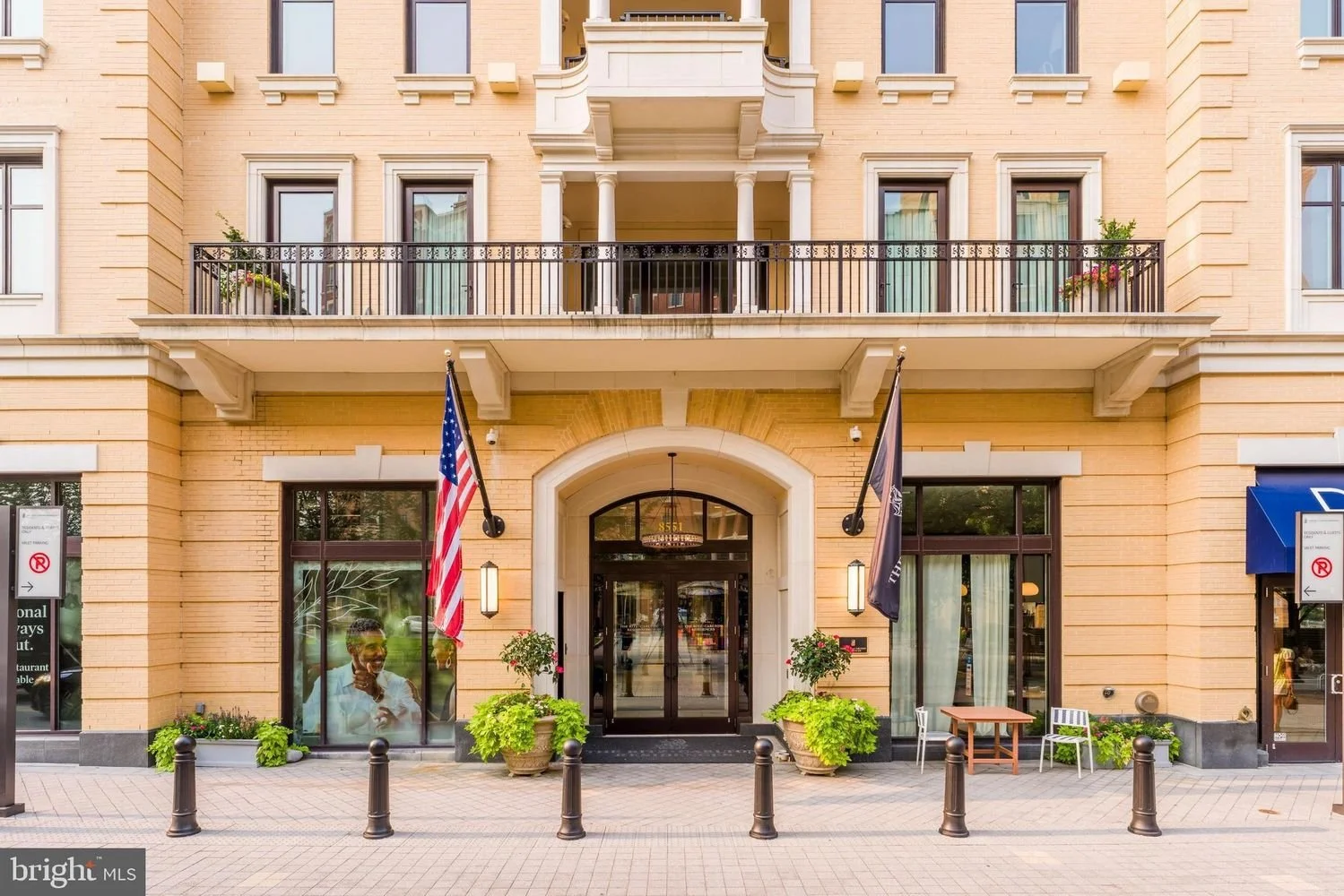 Entrance of a beige brick apartment building with a black door, American flag on the left, and another flag on the right side. There are potted plants and a woman inside looking out through the ground-floor window. A small outdoor seating area with a table and two chairs is on the right. Black bollards line the sidewalk in front.