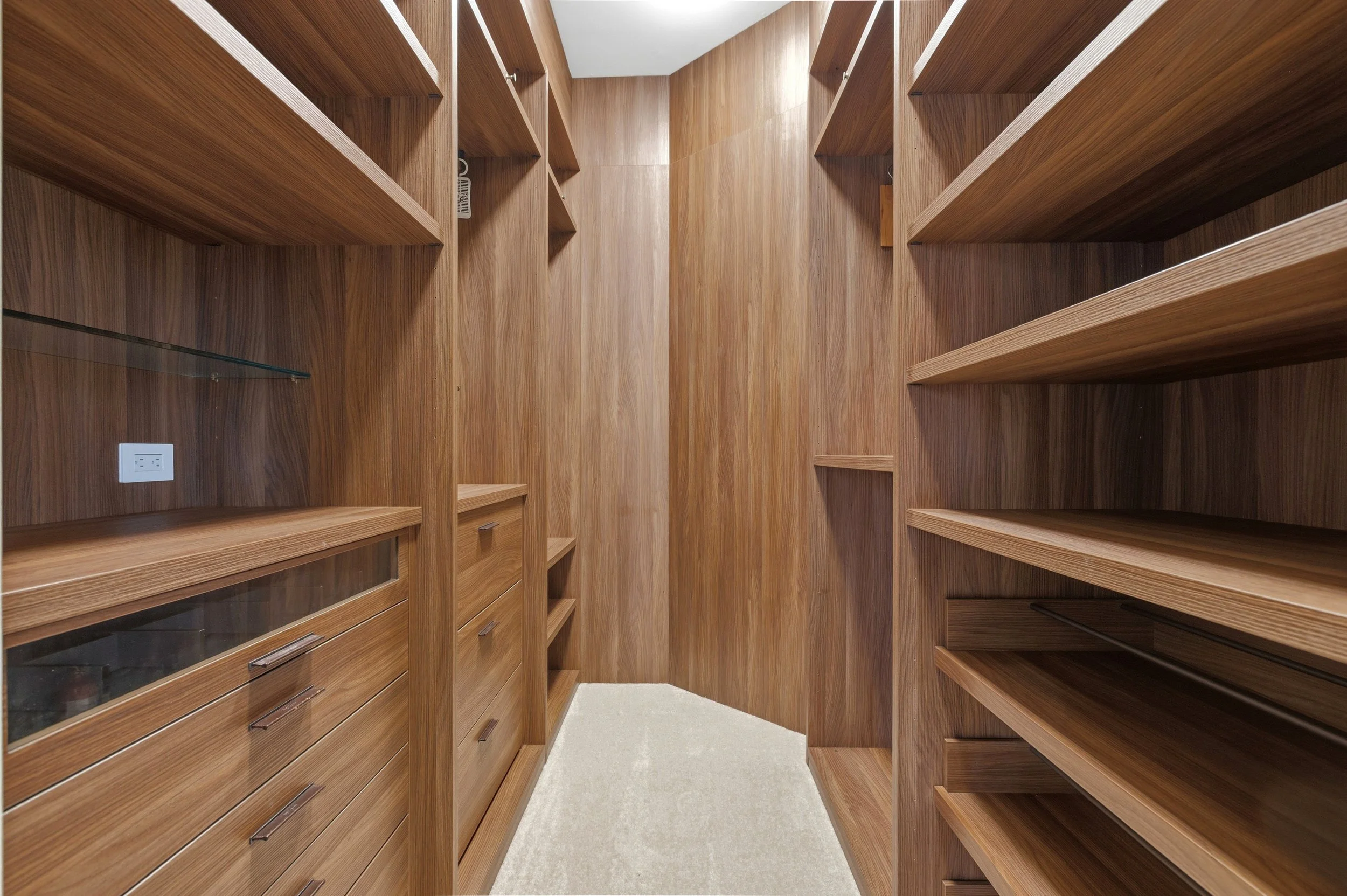 Walk-in closet with wooden shelves and drawers, beige carpet, and an electrical outlet on the wall.