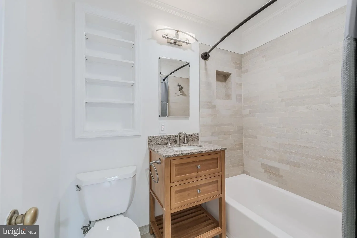 Small bathroom with a toilet, a wooden vanity with a granite countertop, a mirror, a wall-mounted light fixture, and a bathtub with a shower curtain rod. The walls are painted white, and a small recessed niche is in the shower area.